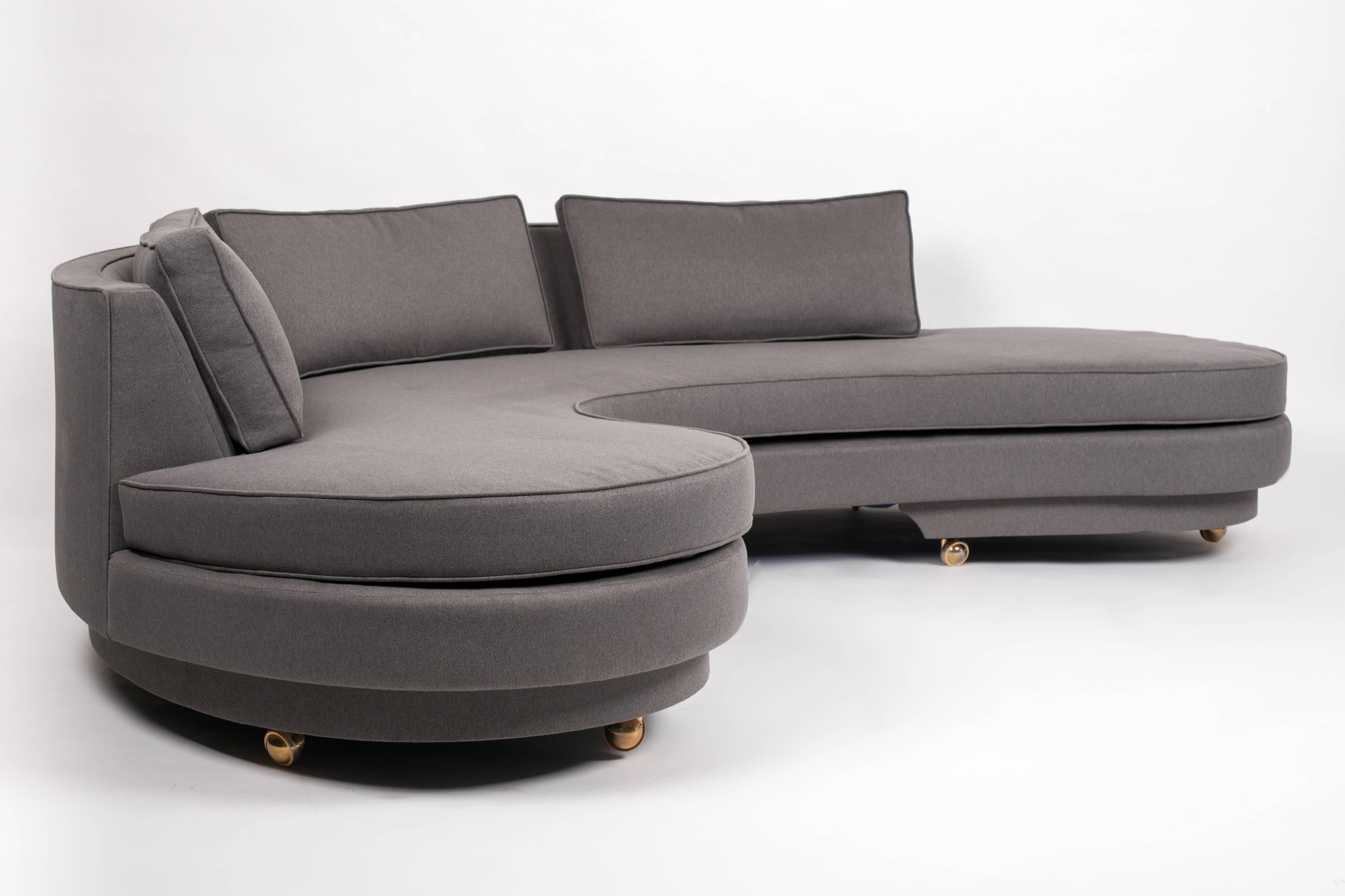 An American-made, semi-circular sofa in dark gray upholstery on brass casters. The sofa has an apostrophe-shaped back panel curved around a deep seat with rounded ends on an upholstered base. The seat and back cushions are removable and the cases