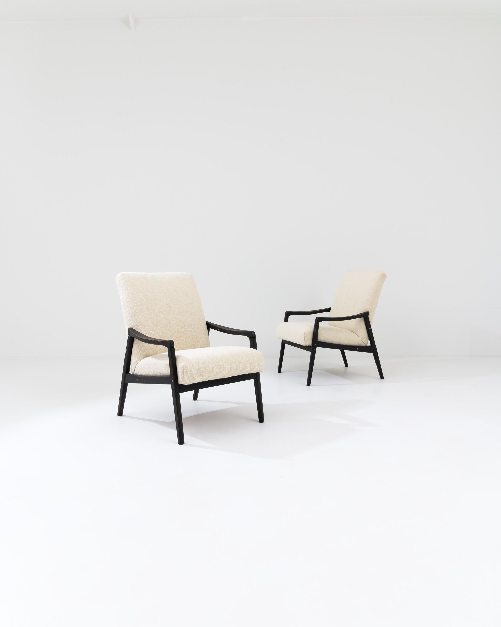 Designed in 1950s Czechia, this pair of exquisite mid-century modern armchairs flaunts sleek silhouettes shaped by the captivating contours of their black-patinated wooden bases with splayed legs and gently curved arms. The white bouclé upholstery