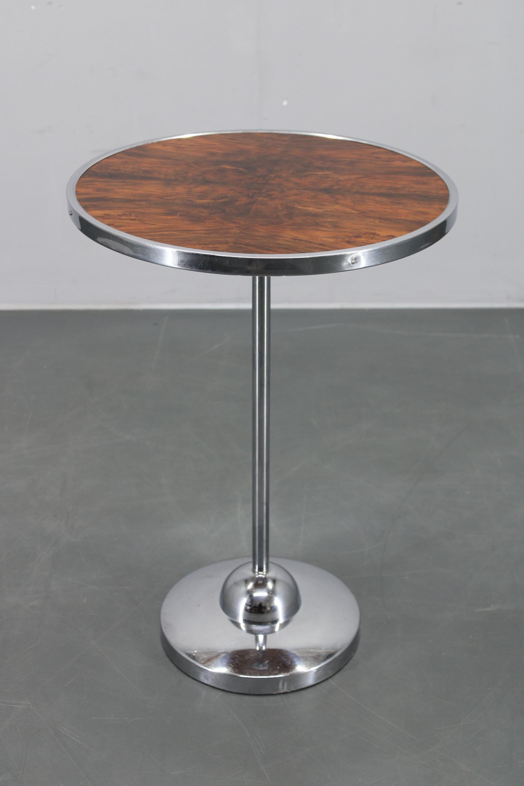 Mesa auxiliar restaurada procedente de Checoslovaquia, alrededor de los años 50, con tablero redondo de chapa de nogal ribeteado en cromo y pata central de pedestal cromada. 
Esta pieza muestra líneas limpias de mediados de siglo y proporciones