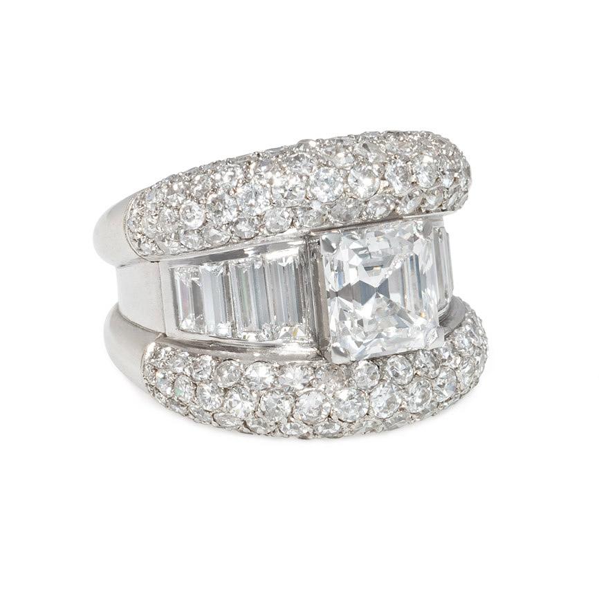 A mid-century diamond ring designed as two curved pavé diamond sections bisected by a line of graduated baguettes and centering on an Asscher cut diamond, in platinum.  French import mark.  Atw 5.79 cts. (atw Asscher 2.43 cts.).  Superb style and