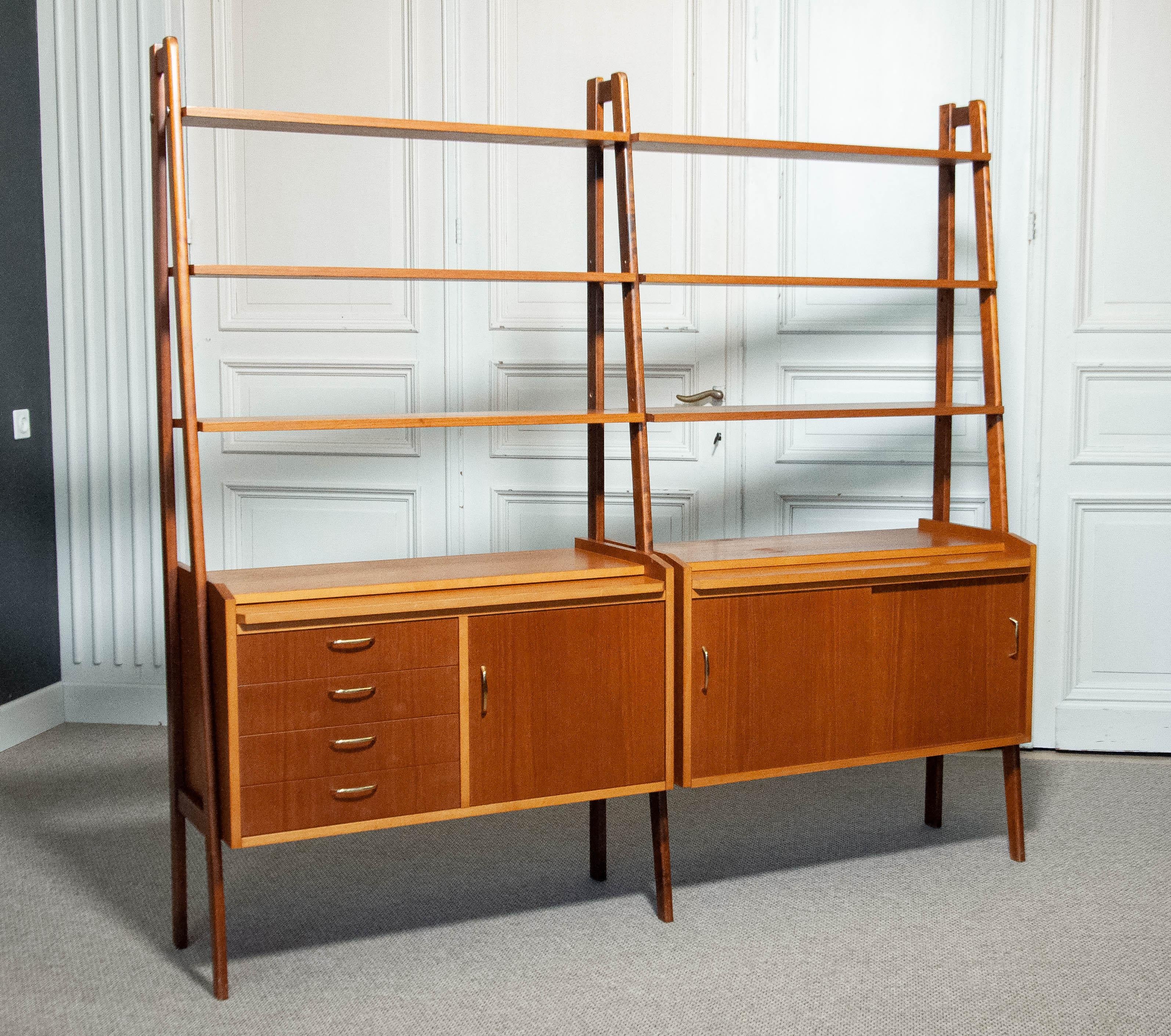 1950s, Sweden — elegant double wall unit crafted in teak and golden teak veneer, with uprights in solid beech.
The composition features two lower cabinets: one with four drawers, a hinged door, and a pull-out writing surface, and another with two
