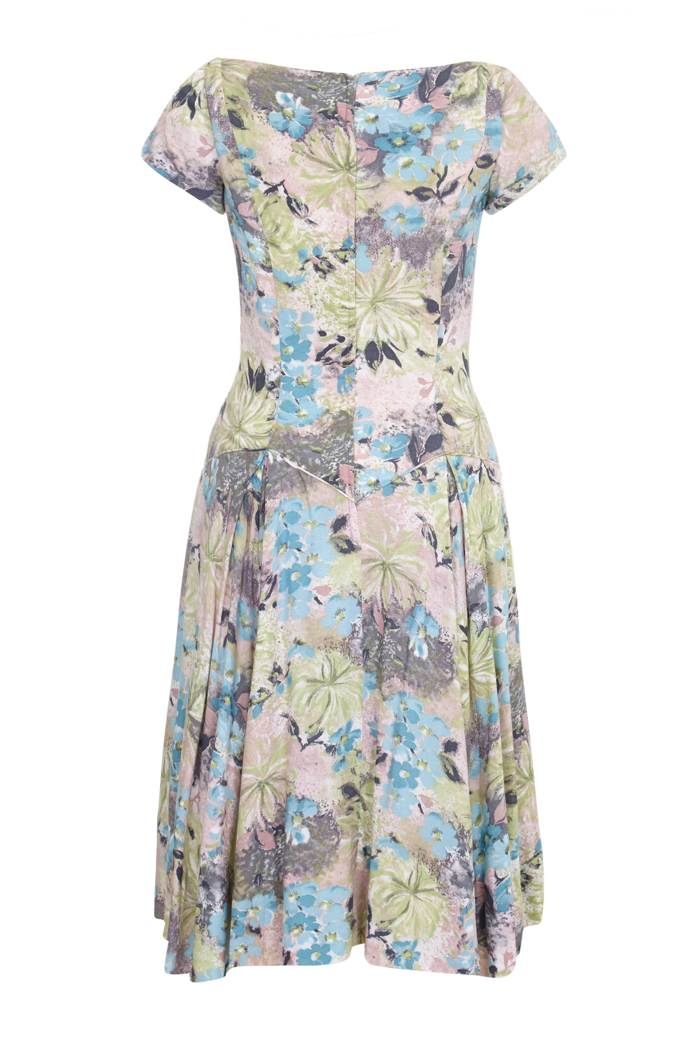 This charming 1950s cotton sun dress with a softly abstracted floral print in soft pastel shades in pink, blue, green and grey is a lovely piece for summer. It features a dropped waist with piping and V shaping to the front and back. There are