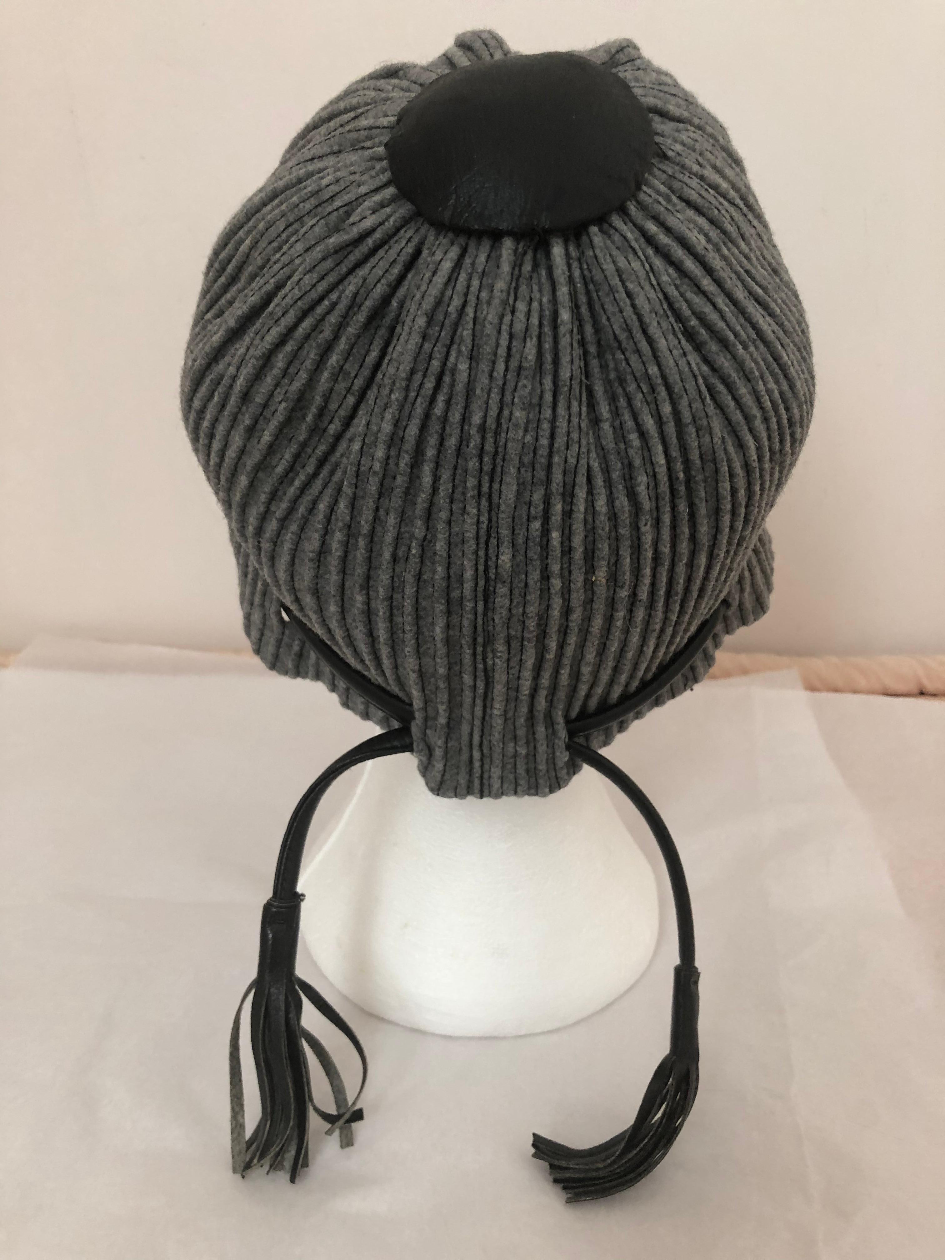 This is a very unique hat from Elsa Schiaparelli, both in style and material. It is made of grey  wool stitched raised ribbing, and black leather lacing ending in tassels. It also has a black leather disc at the top, with gros grain patch on the