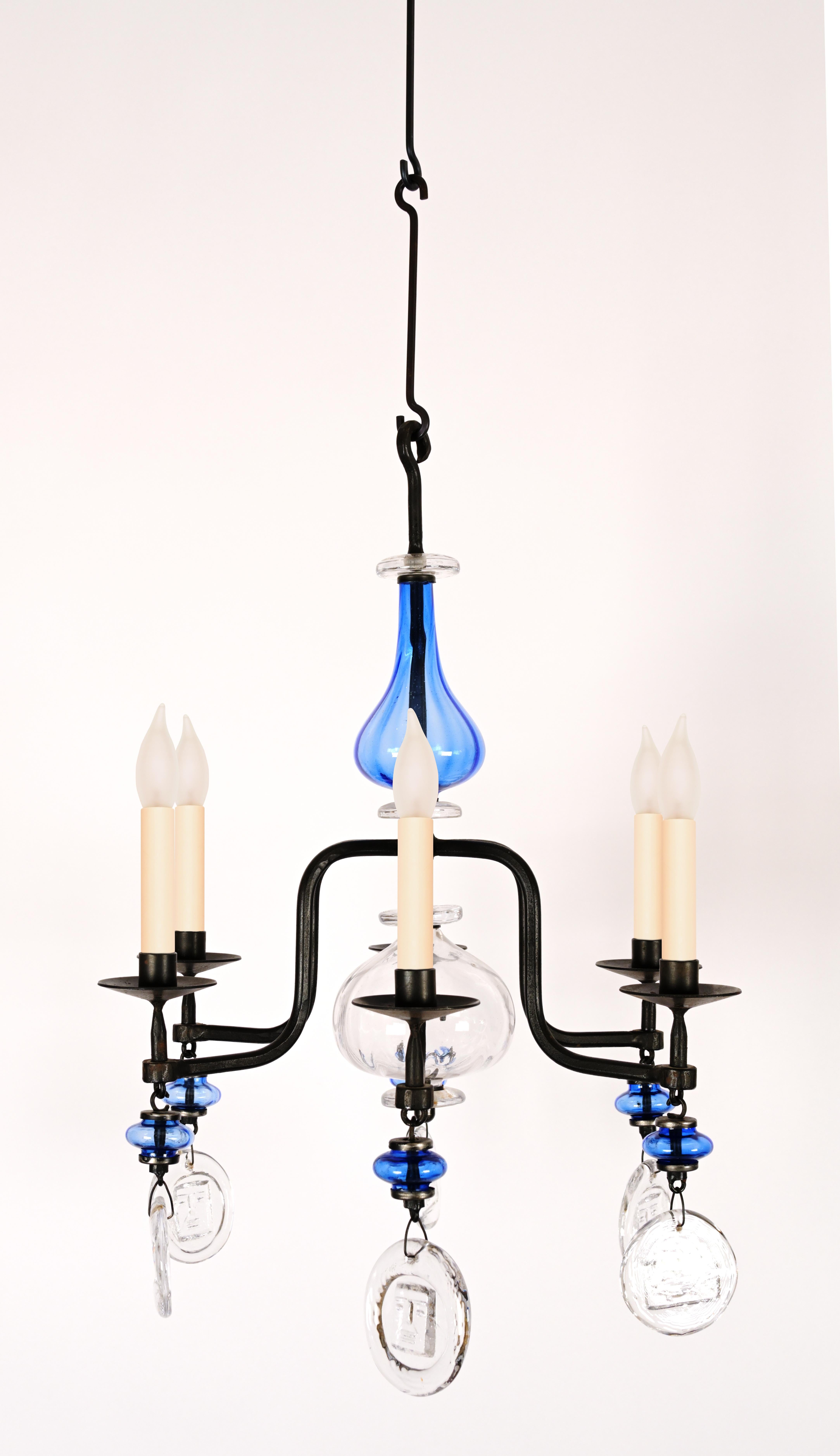 A glass and iron chandelier by Swedish Designer Erik Hoglund (1932-1998) having a bulbous blue blown glass center with 6 curved arms decorated with circular glass chips with imprints of playful faces and symbols. Includes standard wiring.