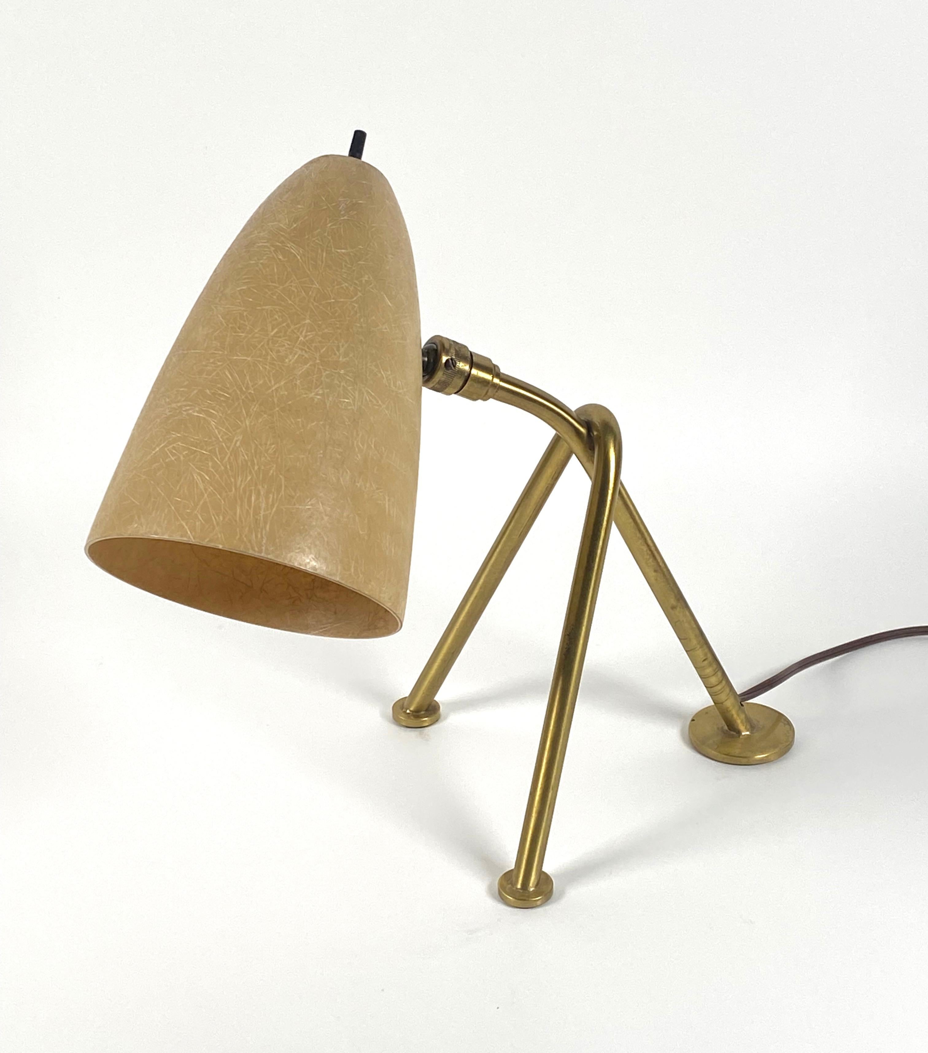 Small table lamp crafted of brass and fiberglass, having a conical fiberglass shade in a soft gold color compilmentary to the solid brass three legged grasshopper form base with round pad feet the quality of construction is outstanding along with a