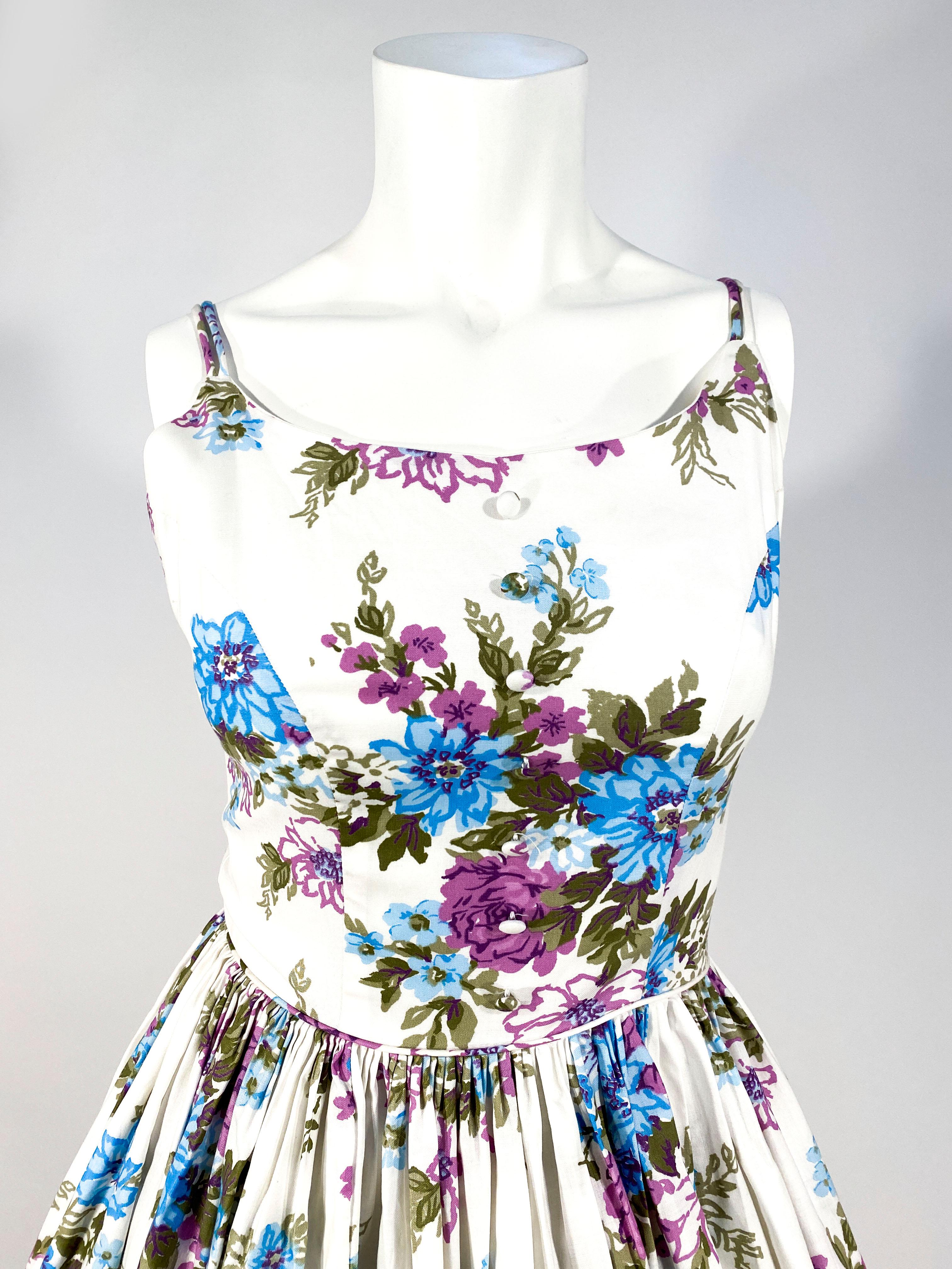 1950s Handmade cotton floral printed day dress featuring tones of aqua, purples and greens on a white field. The torso is fitted to a very full pleated skirt starting at the waist. The bodice has hand-covered button decorations the is lined in the