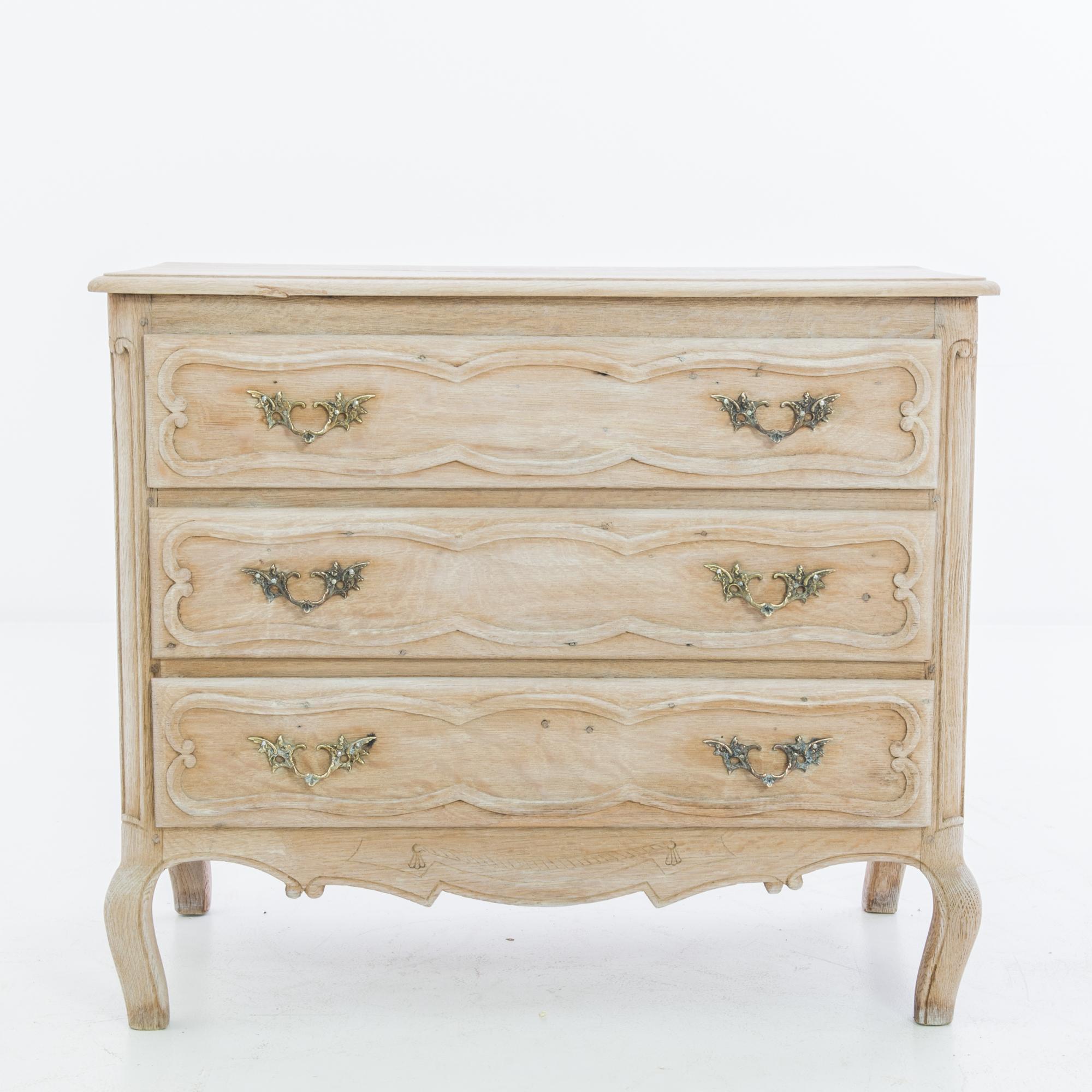 A three drawer oak chest from France, circa 1950. The wood has been restored in our workshops to a pale finish, revealing the natural rose tone of the oak. A lively pattern of carved scrolls dances across the apron; cabriole feet lend elegant