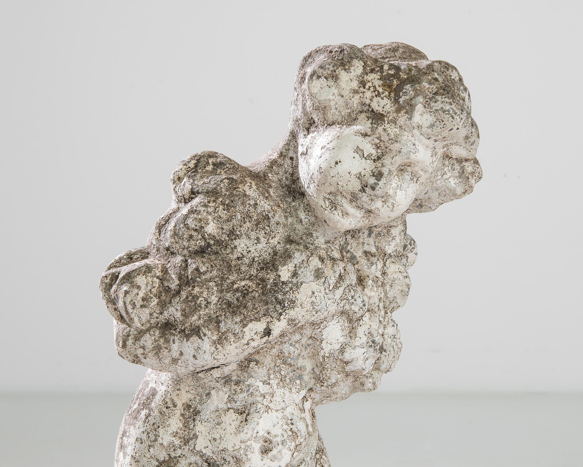 A concrete statue from 1950s France, depicting a cherub holding a garland of flowers. An aged patina dapples the surface of the concrete, inflecting the original white painted finish with notes of lichen and stone. The graceful pose and soft outline