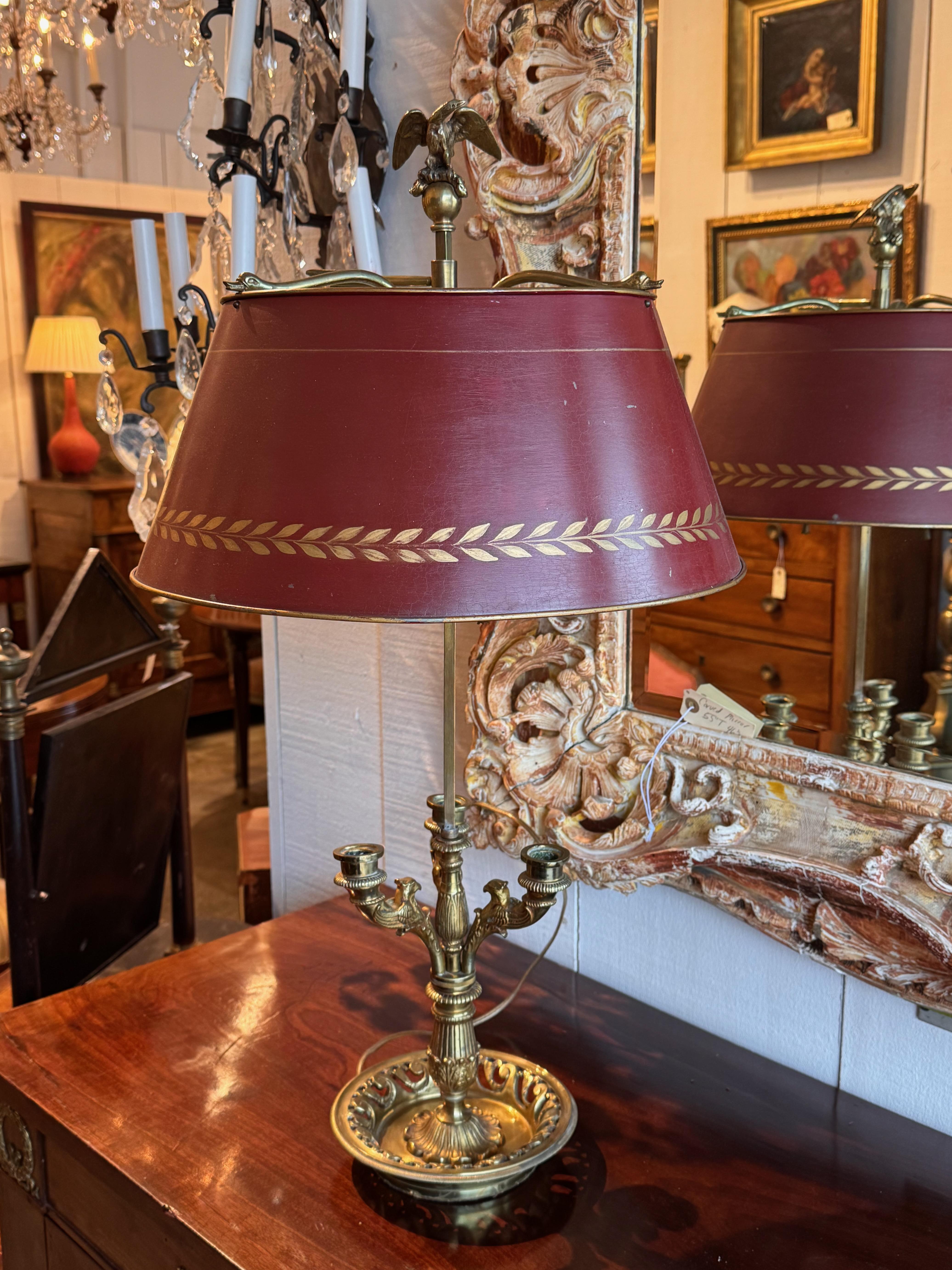 Elegante lámpara bouillotte de estilo Imperio francés, con una base de latón con detalles clásicos y un trío de brazos estilo vela, cada uno adornado con cabezas de águila finamente fundidas. La pantalla de tole ajustable está acabada en rojo