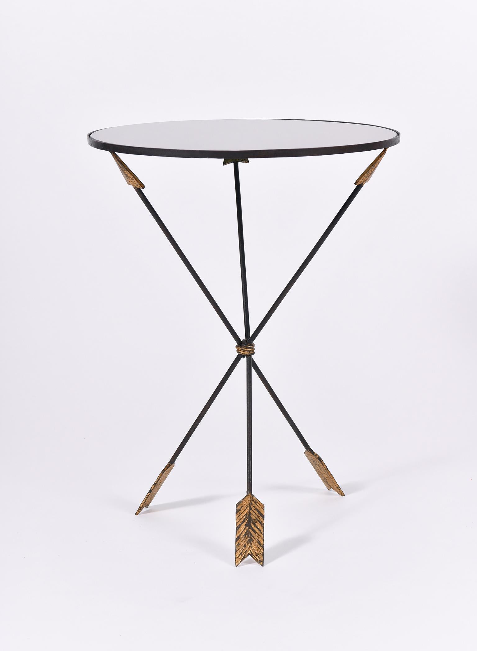 A gilt and black enameled iron occasional table, the arrow shaped tripod supporting a smoked mirror glass circular top
France, circa 1950.