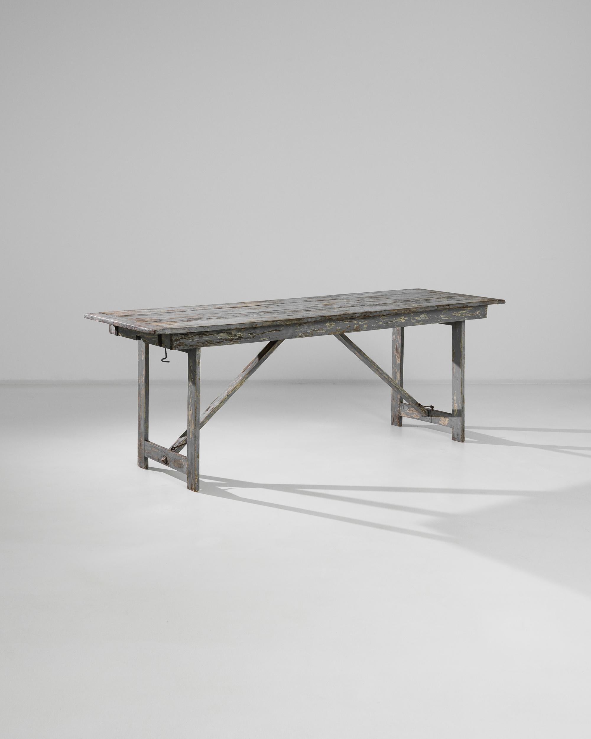 A wooden table from 1950s France with a rustic patina. The original coat of dove-grey paint has weathered over time, revealing notes of primrose yellow and glimpses of the natural wood beneath. Diagonal struts lend a note of complexity to the