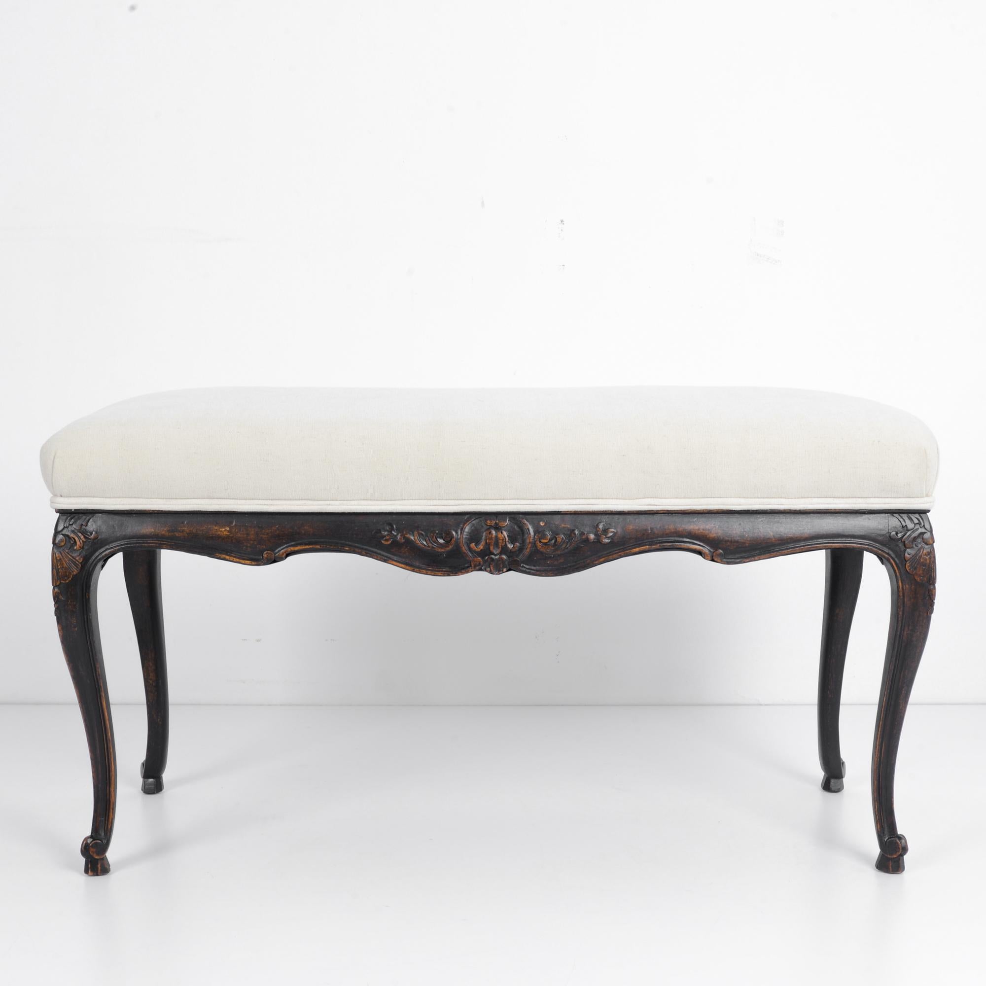 This wooden bench was made in France, circa 1950. The ivory white upholstered and well-padded seat brings out the intense shade and beautiful patina of the wood. The curves of its cabriole legs, scalloped apron, and intricate floral carvings add