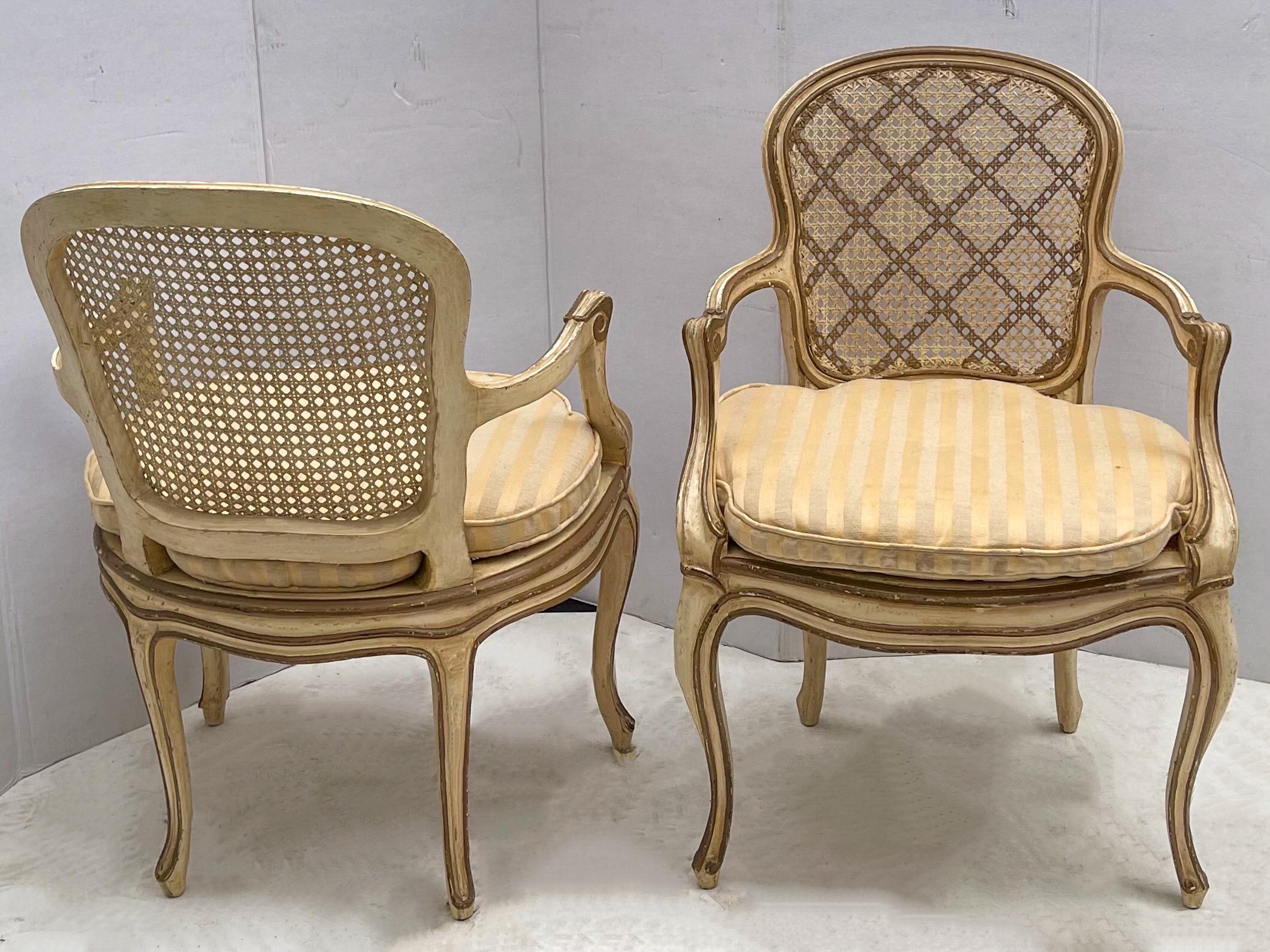 This is a lovely pair of 1950s ivory and gilt painted French bergere chairs. They are in very good condition with some age appropriate patina. The silk striped cushions, however, are vintage and show wear.