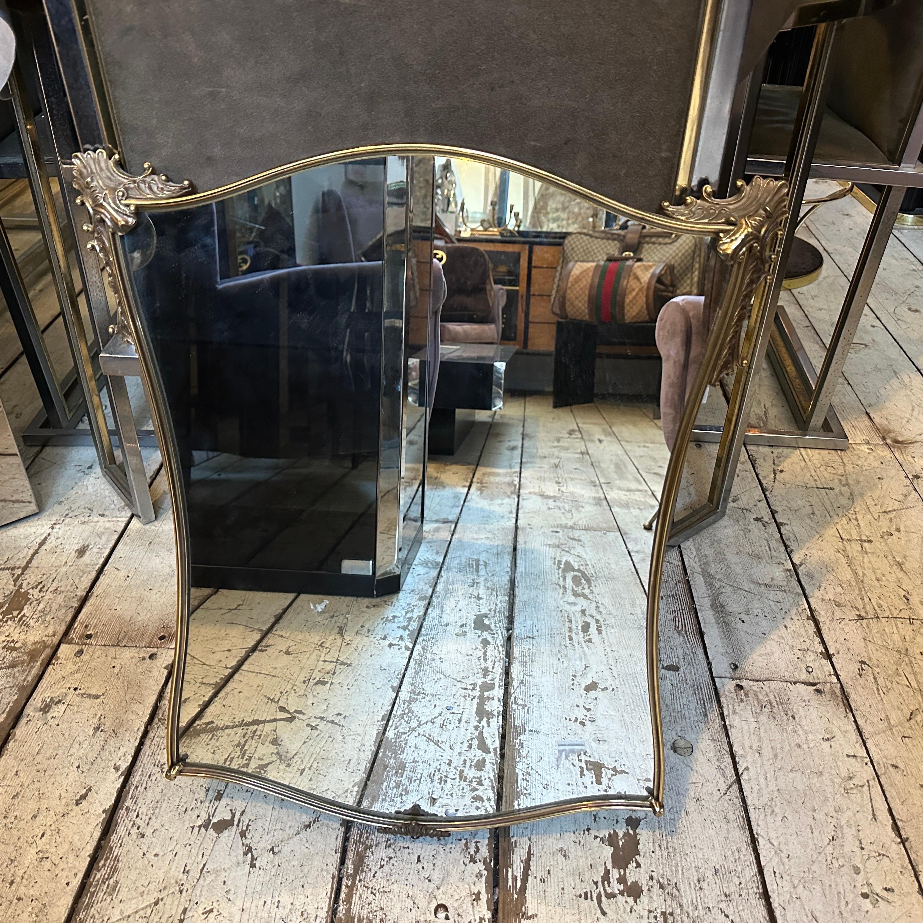 Le miroir présente une silhouette élégante et subtilement curviligne, plus étroite à la base et s'élargissant légèrement au sommet, ce qui lui confère une forme gracieuse et féminine. Le cadre en laiton est fin et délicat, soulignant le minimalisme