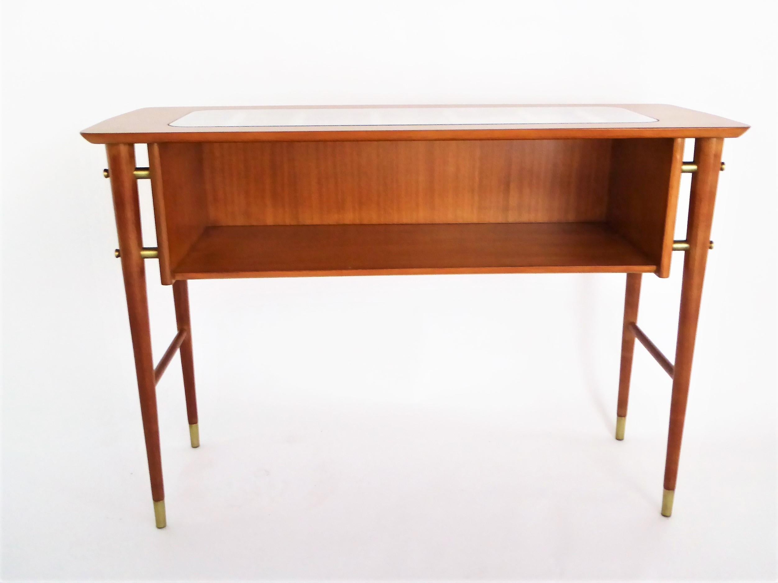 A 1950s Gio Ponti style Petite console table with shelf in blond mahogany with brass sabots and side mounts. Featuring an inset glass on top with slat supports and stylized screw-top wood inlays. Restored finish with original glass with minor age