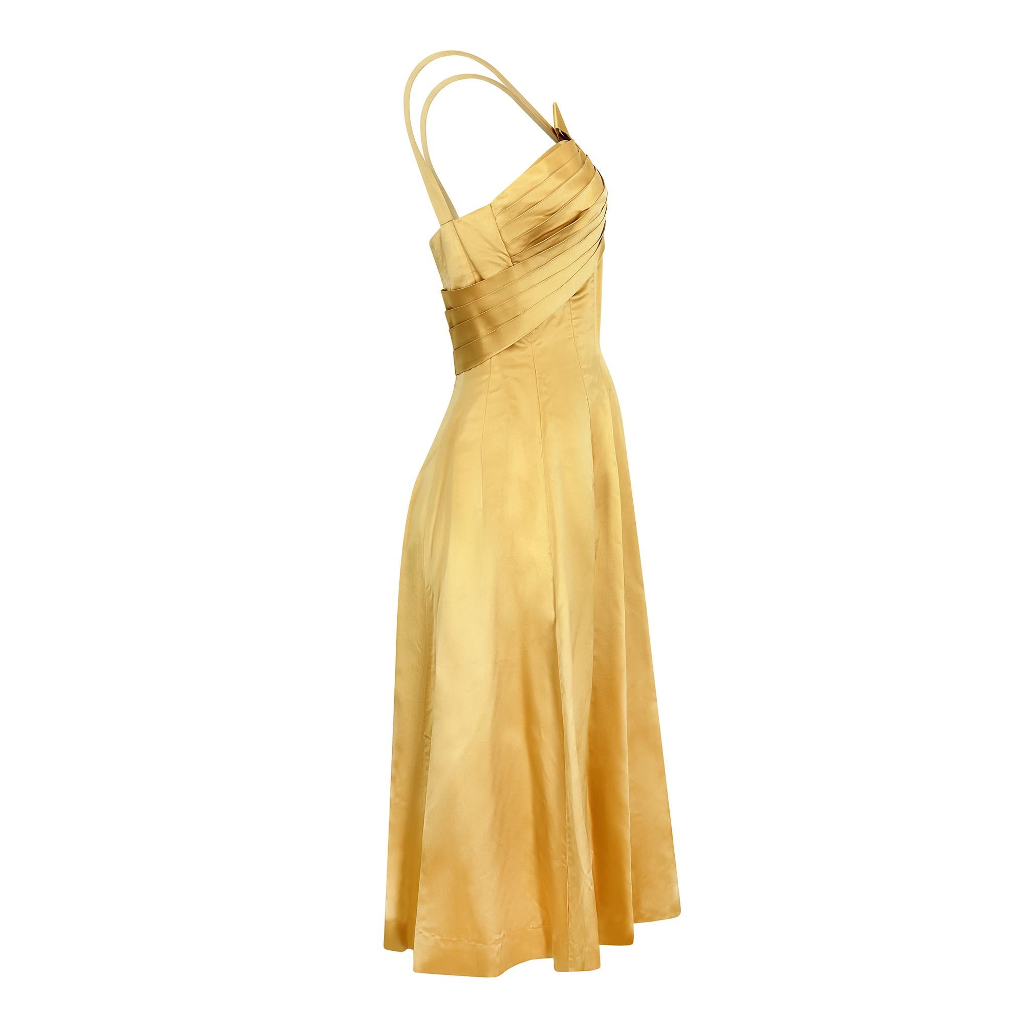 A highly desirable, original, 1950s cocktail dress which could easily be worn as an alternative wedding dress. The fabric is a beautiful gold coloured silk satin and the dress features a well executed pleated bodice with an asymmetric bow with long