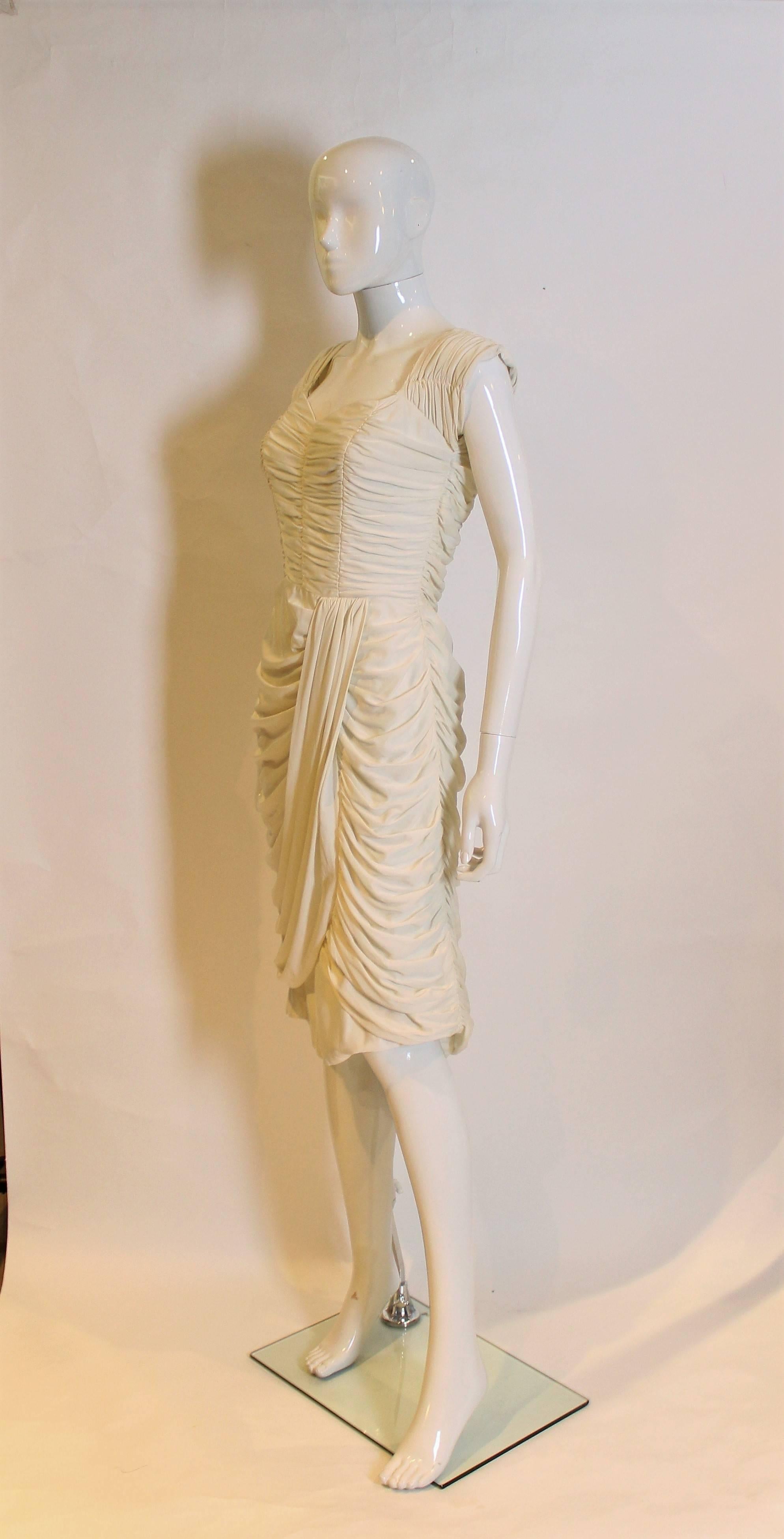 Ein weißes Cocktailkleid mit Raffungen und Falten, vielleicht ein Hochzeitskleid?
Das Kleid hat einen herzförmigen Ausschnitt und einen umgeschlagenen Rock,  zentraler Rückenreißverschluss und ist vollständig gefüttert.