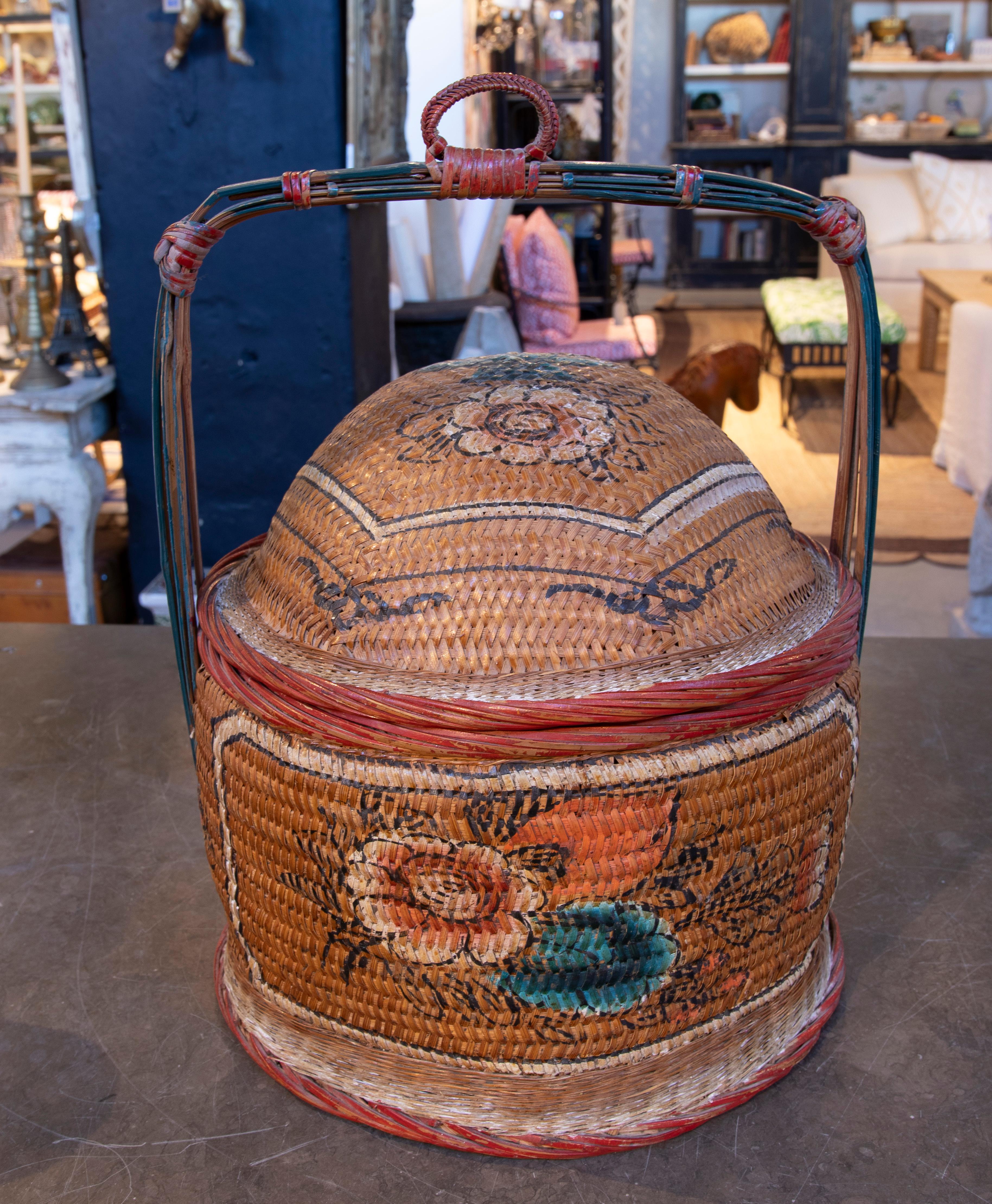 Bellissimo cesto in vimini intrecciato a mano e decorato con dettagli floreali dipinti a mano in tonalità vivaci. Il suo design è caratterizzato da un coperchio a cupola e da un manico rinforzato con fibre colorate, che lo rendono funzionale e