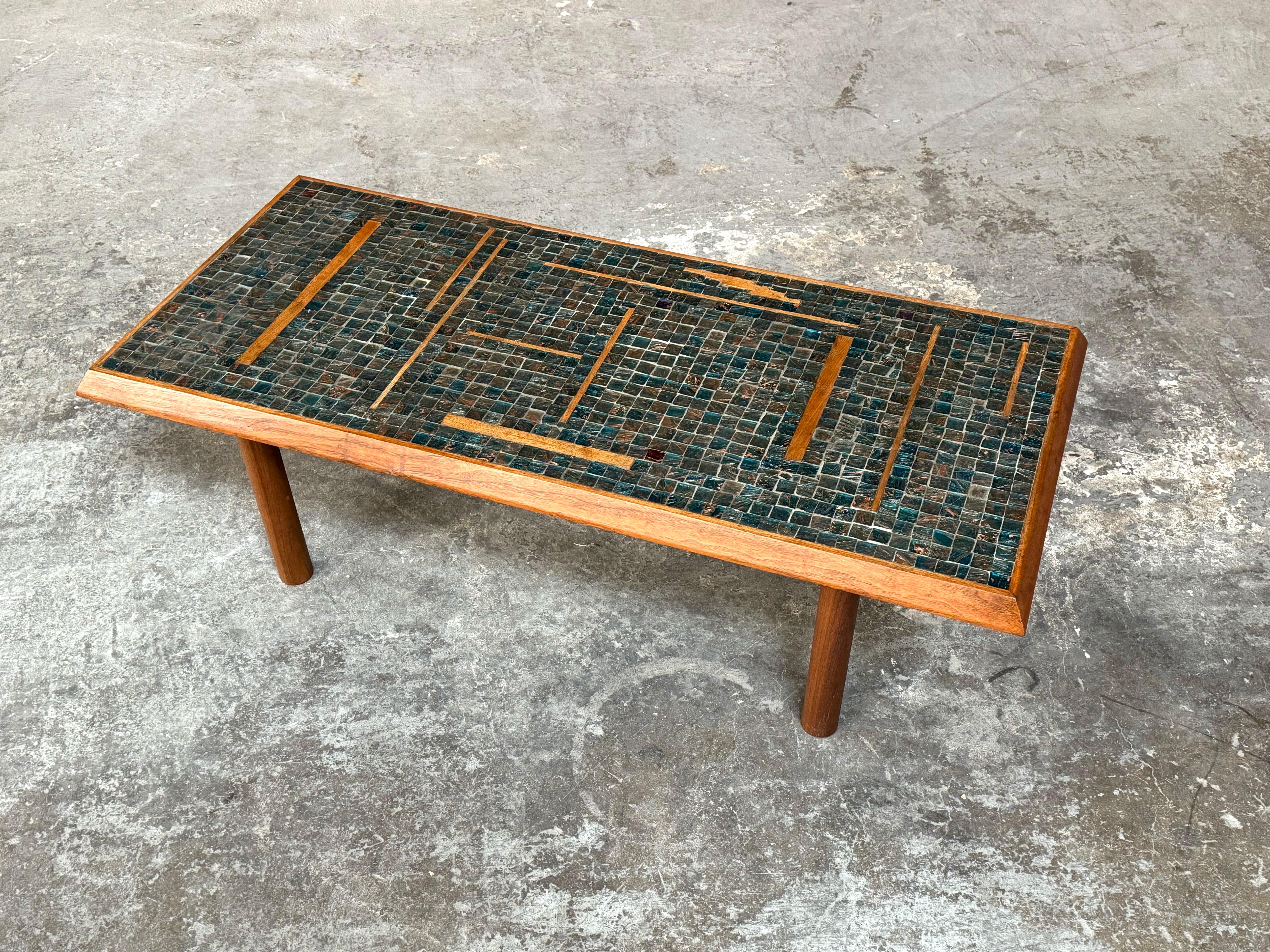 Handmade glass tile mosaic coffee table with walnut inlay and edge trim. A compact size coffee table with earth tone colored tiles in browns and blues with an abstract / geometric pattern walnut inlay, the table legs are a cylindrical walnut. The