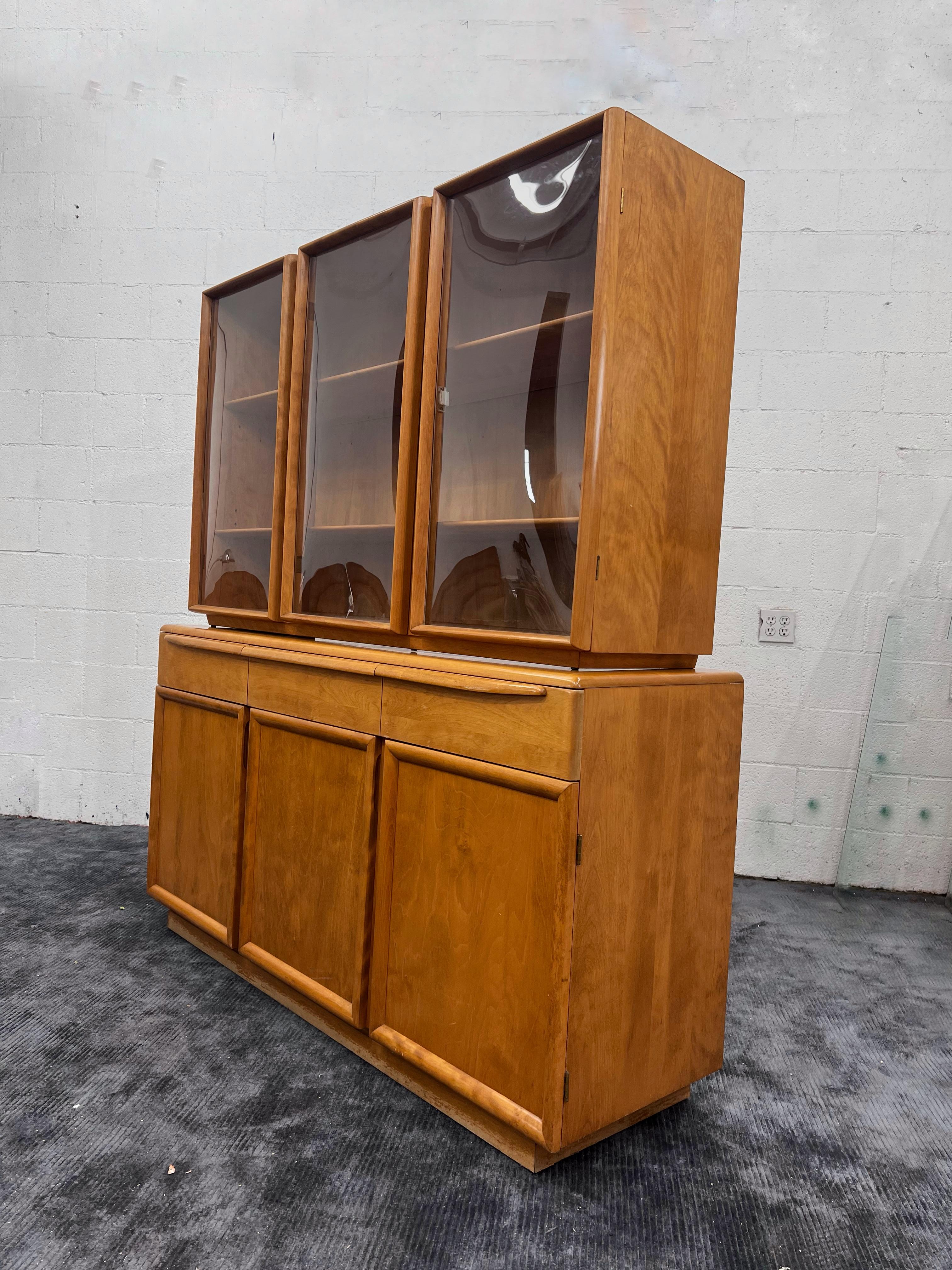 Mid-Century Modern Heywood Wakefield two-part china cabinet, circa 1955. The original finish has aged to a dark amber / tobacco color. 
The top cabinet has three bubbled glass doors that open to reveal shelves. The bottom cabinet has two drawers