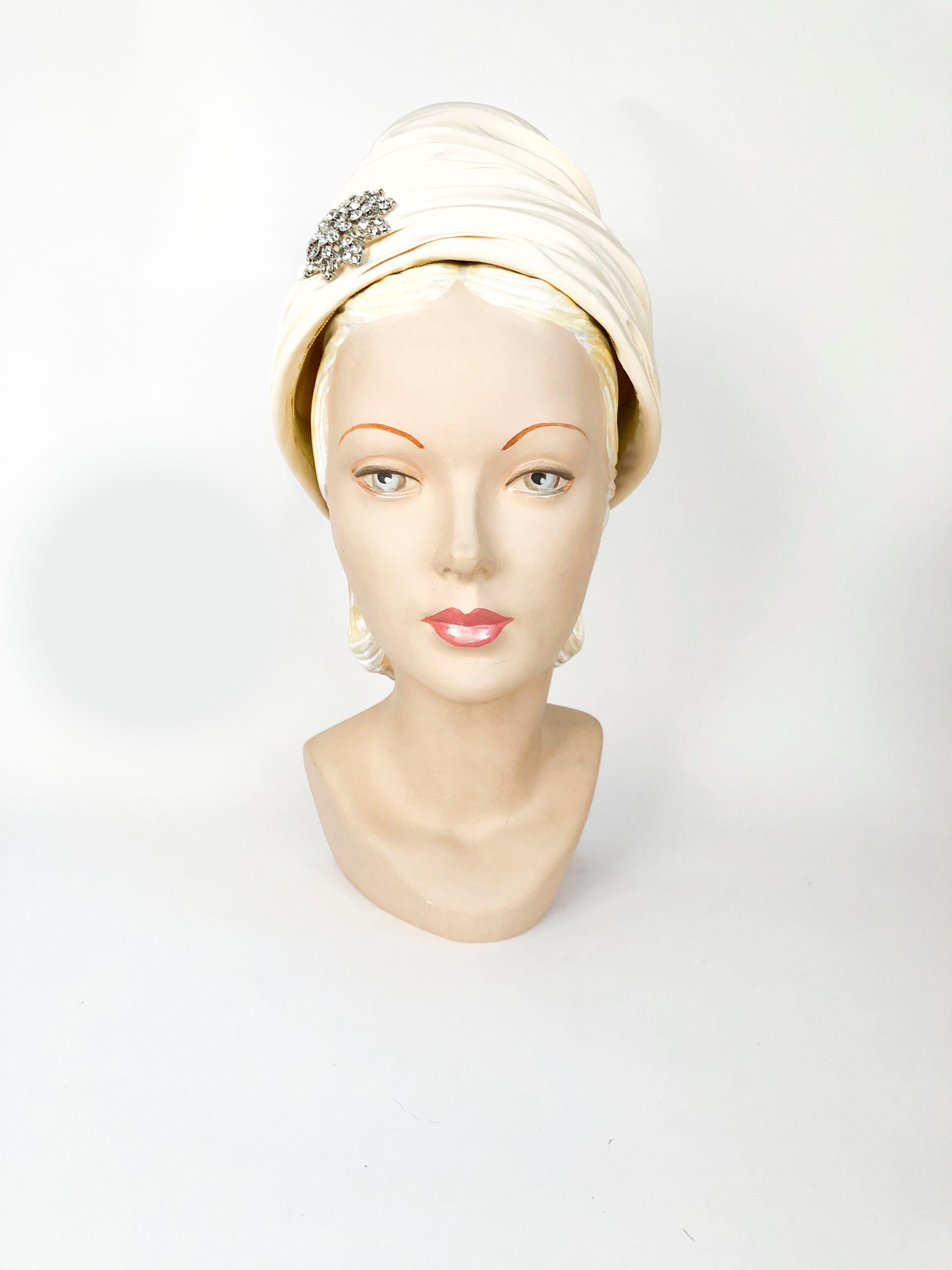 1950s/1960s High-Fashion Structured Turban with a high-crown in cream bone color that is gathered and layered around the entire hat. Adorned with a clear rhinestone accent