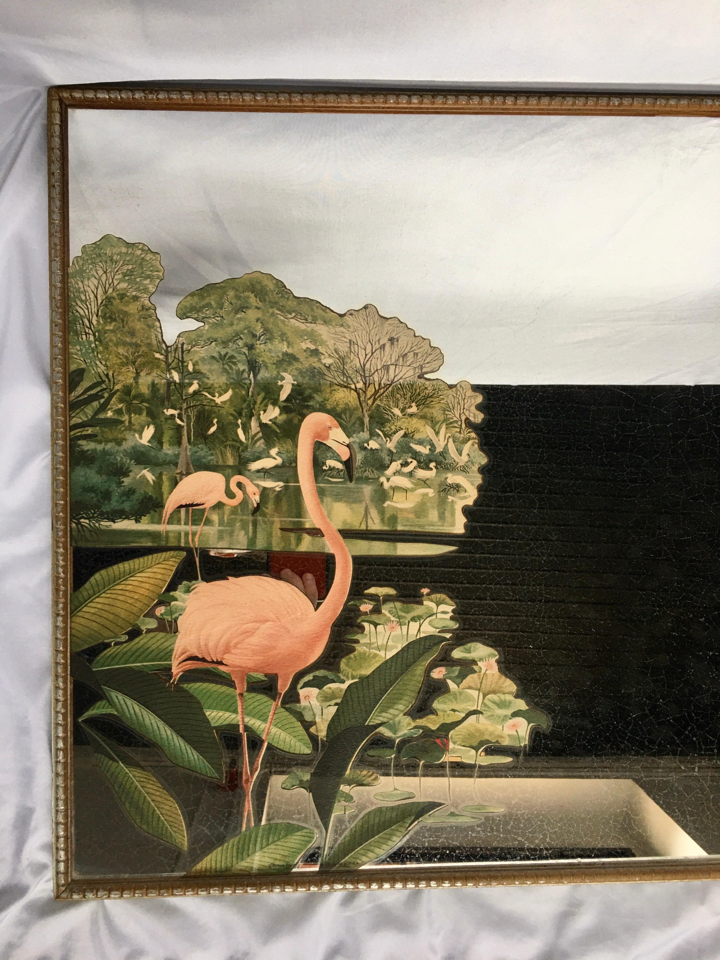 Hollywood Regency decorative wall mirror featuring a painted tropical design of pink flamingos and palm leaves. This midcentury mirrored art piece is framed in a gold/silver tone wood frame, circa mid-20th century. Pair available.
