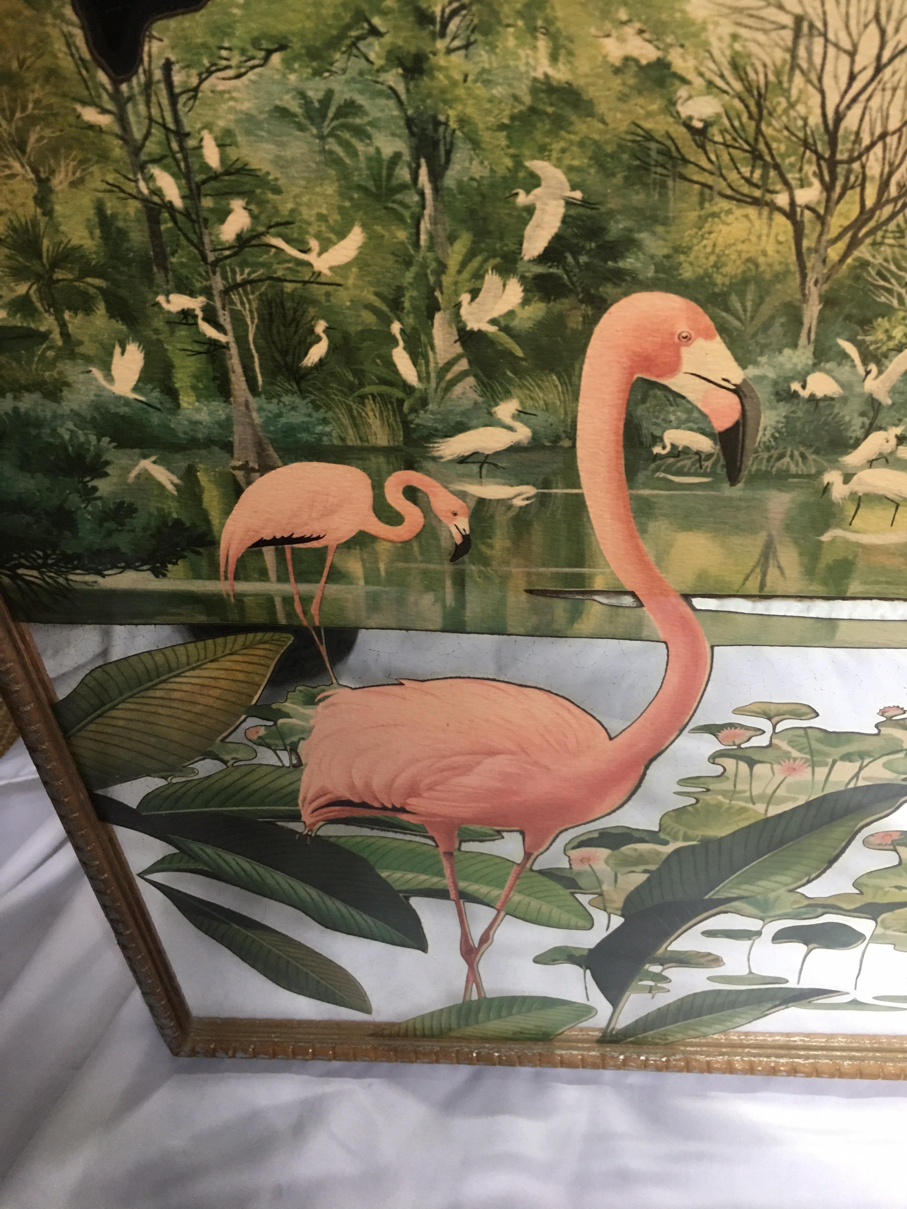 Hollywood Regency decorative wall mirror featuring a painted tropical design of pink flamingos and palm leaves. This midcentury mirrored art piece is framed in a gold/silver tone wood frame, circa mid-20th century. Pair available.