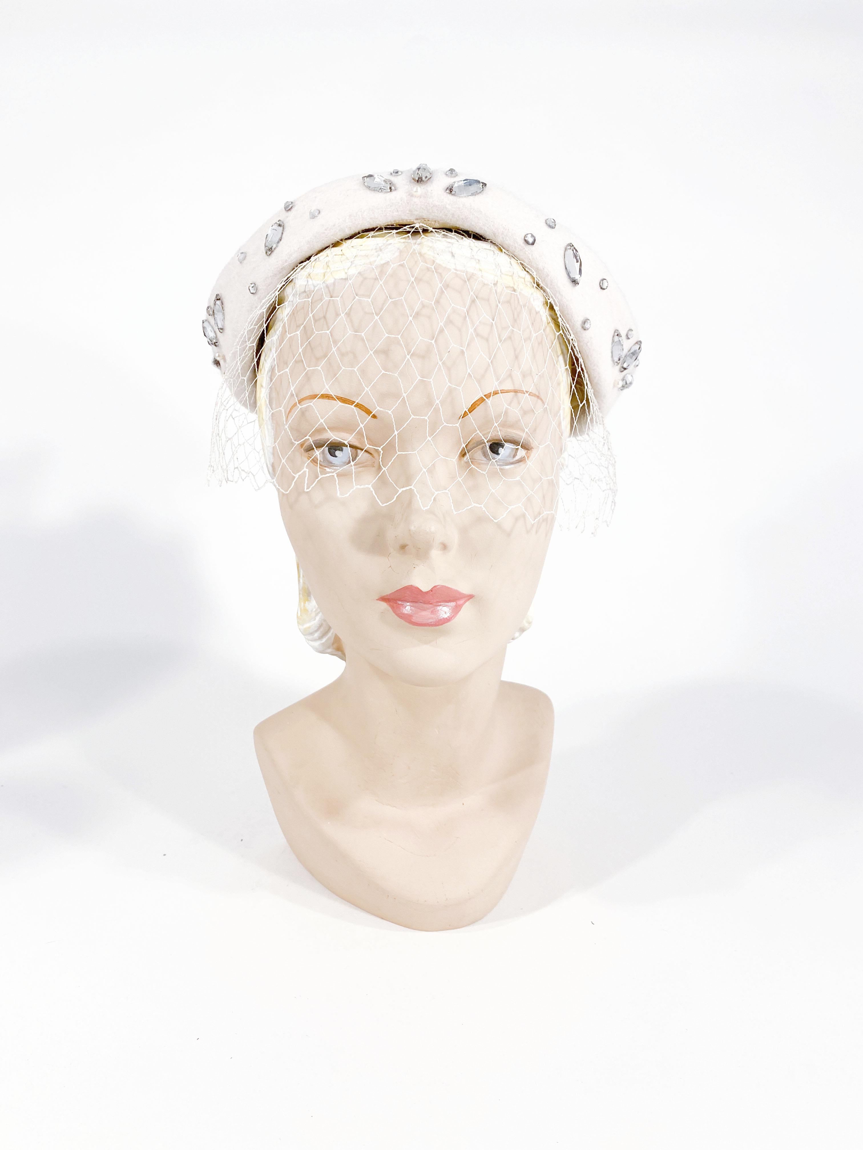 1950s cream-colored fur felt hat with rolled brim decorated with large clear rhinestones and pearls. The matching net is attached in the interior and reaches to the end of the nose. Meant to be worn and secure the hat to the head.