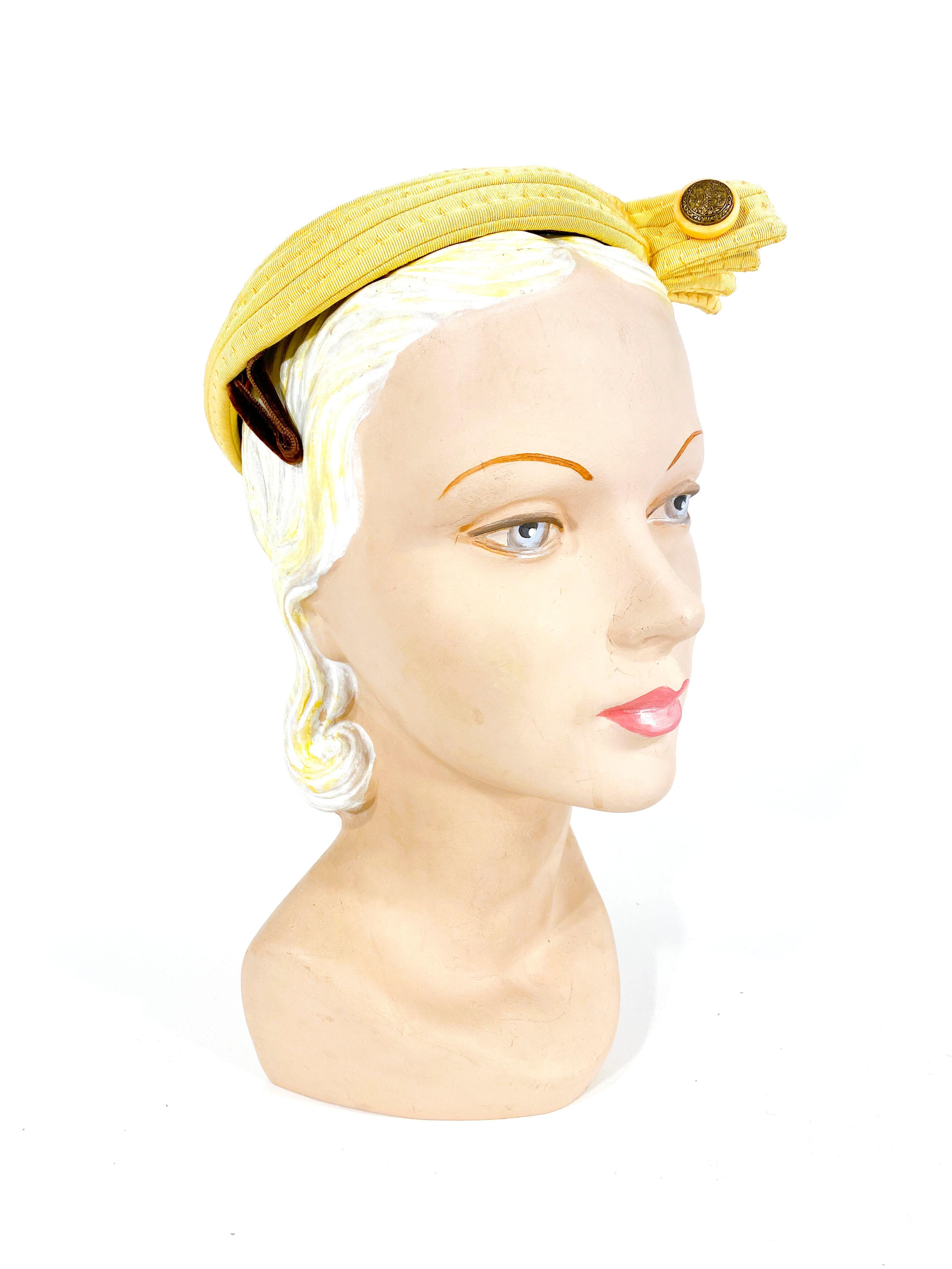 1950s soft yellow hat made of rolled grosgrain in a snail pattern finishing at the top. Each row of grosgrain is topstitched and the front of the hat an off centered wing adorned with an etched brass button. The frame of this hat is wired for shape
