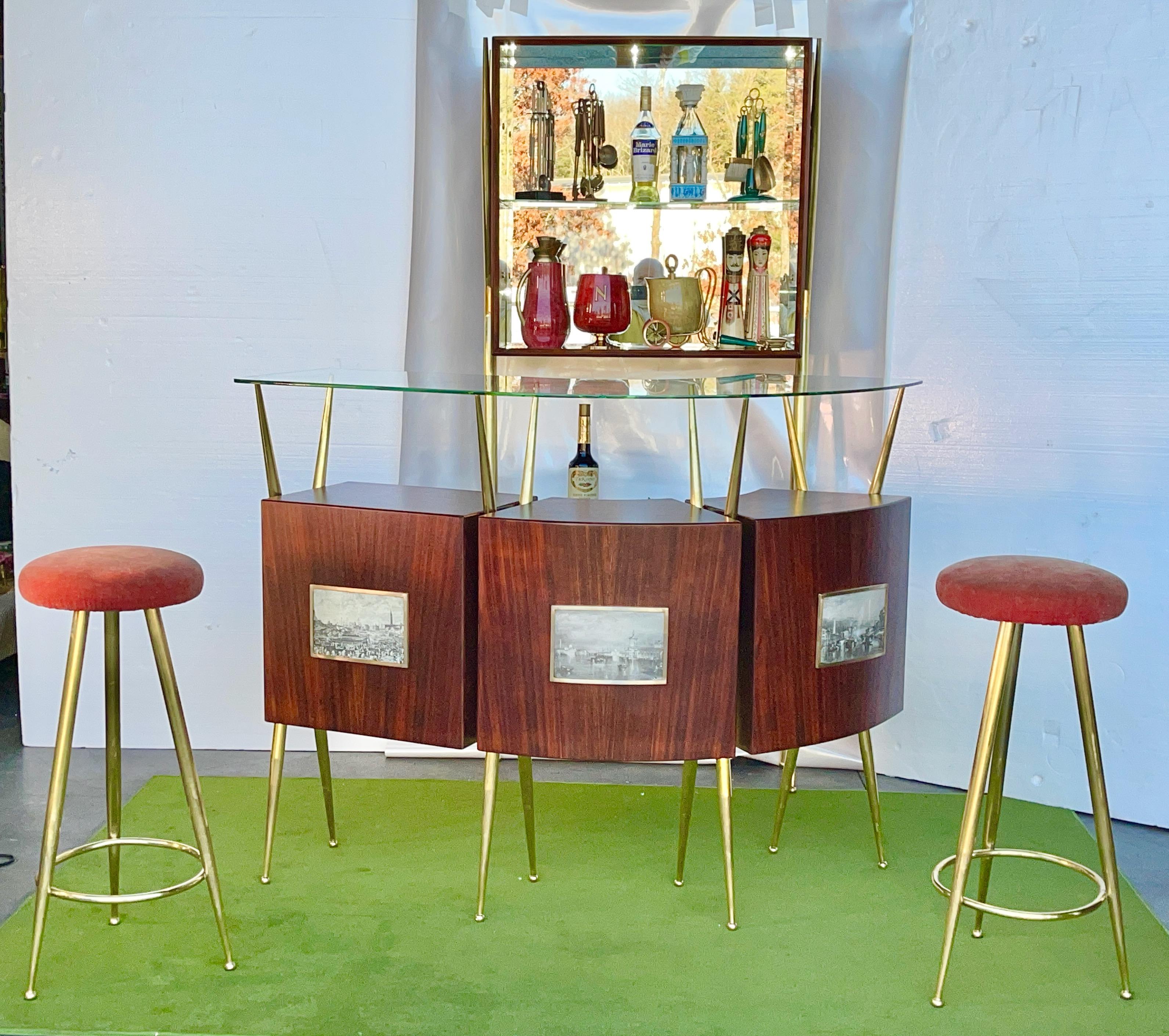 1950's Italian standing bar, back bar and pair of brass three legged bar stools.
Standing bar has three open cabinets in satin finished palisander veneer held aloft by angular tapered tubular brass scaffolds. and a floating curved glass