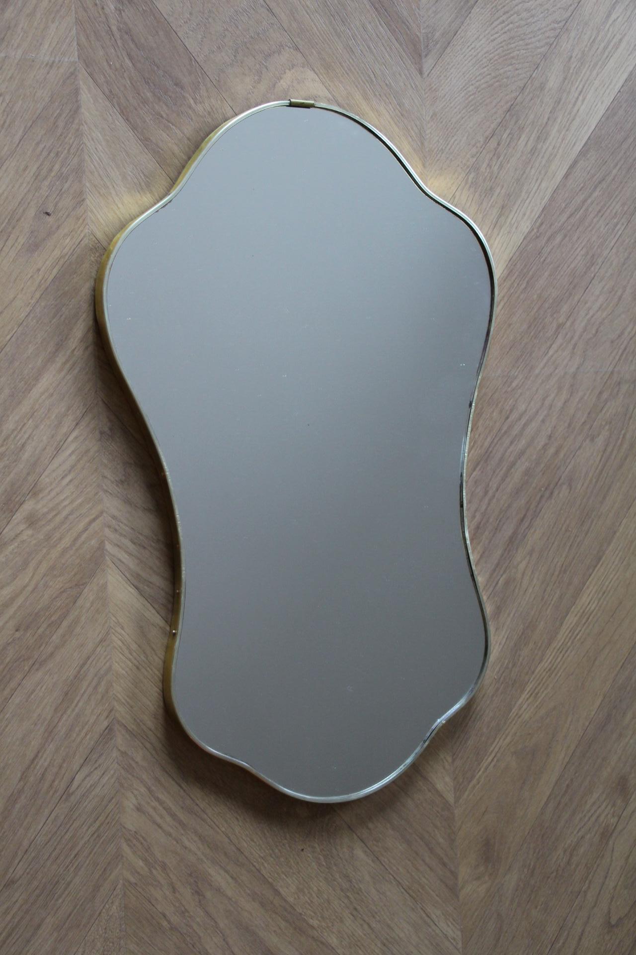 This piece is an elegant Italian wall hung brass mirror with a tapered silhouette, slim line brass edge and soft radial corners. It has exaggerated curves that give it a lot of character. The curved silhouette is quintessential of Italian 1950s