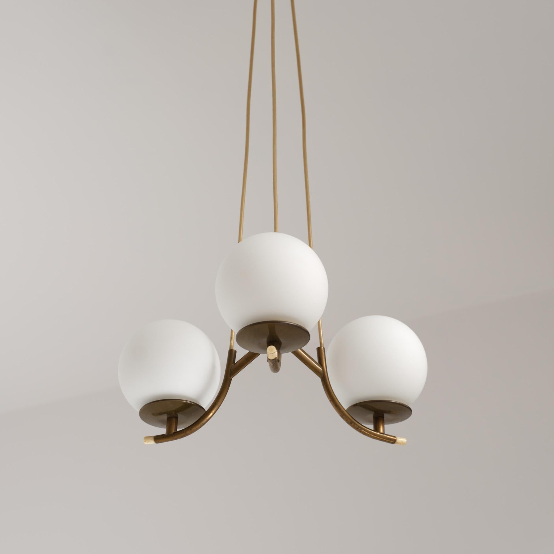 Italian chandelier from the 1950s, composed of a brass structure with three opaline glass shades.
Each shade is positioned on a curved brass arm, arranged around a central joint that defines the overall geometry. The brass shows a natural patina