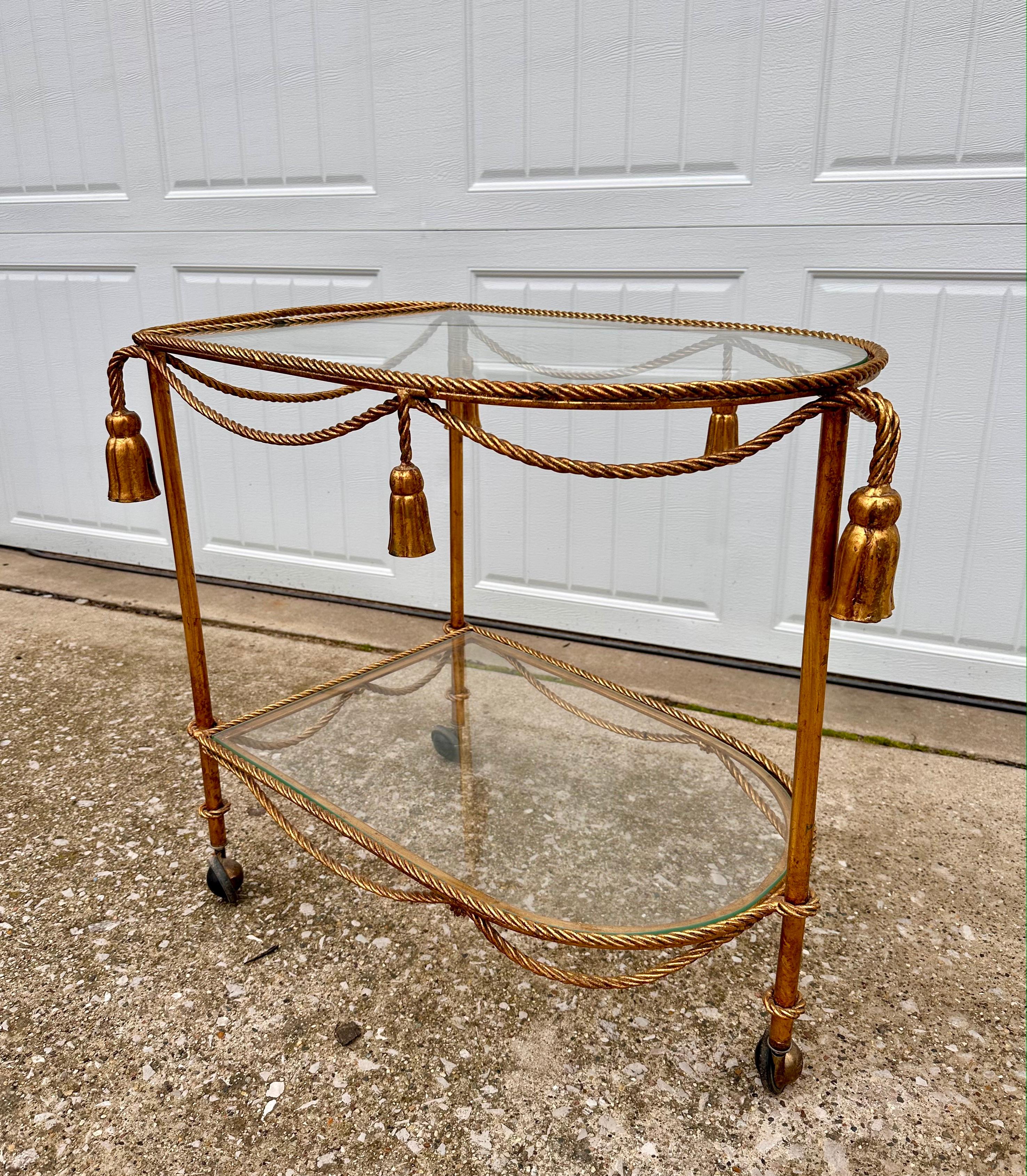 Vintage Hollywood Regency gilded two tiered tasseled bar cart on wheels. In great condition no chips or cracks in the glass, wheels roll smoothly and minimal patina on cart. Some patina and chipping due to age. Some very light scratches on the