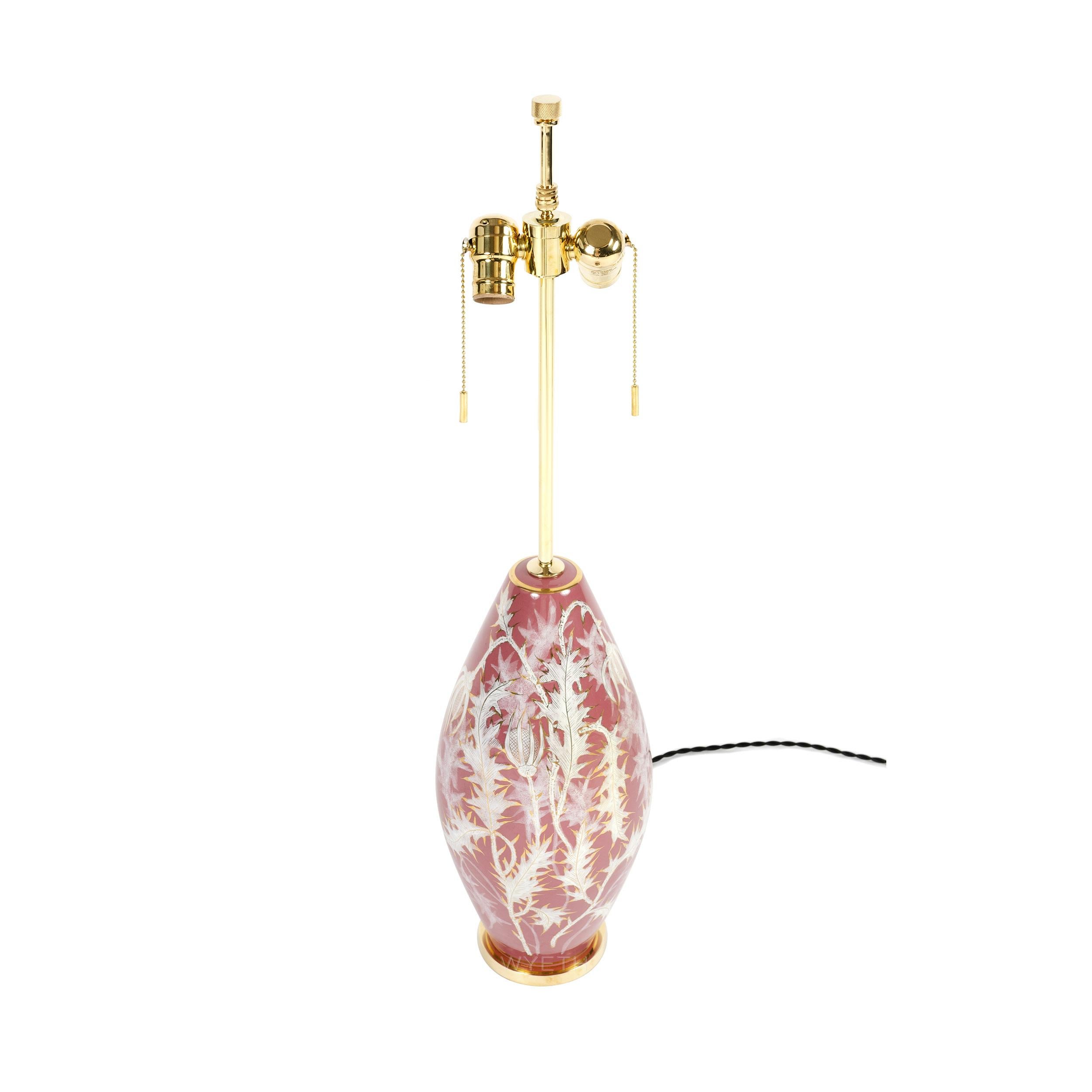 Ceramic table lamp with an ovoid shaped body, hand decorated with white thistle leaves and vine with gold accents on a light, coral colored background.