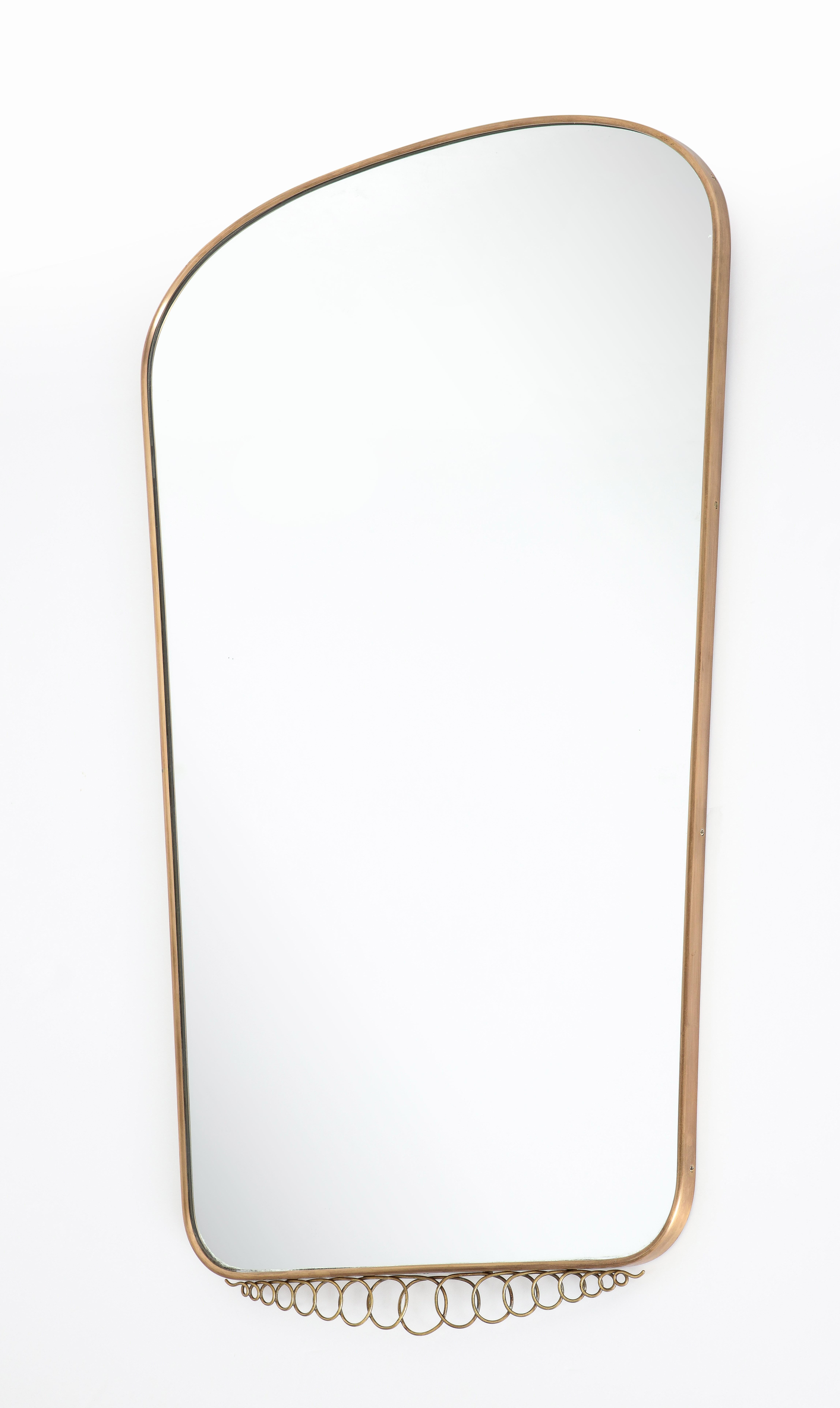 Miroir mural moderniste italien des années 1950 en laiton de grande forme avec un sommet arqué qui se rétrécit vers le bas avec un joli élément décoratif en laiton attaché. Cet élégant miroir du milieu du siècle illustre dans cette monture