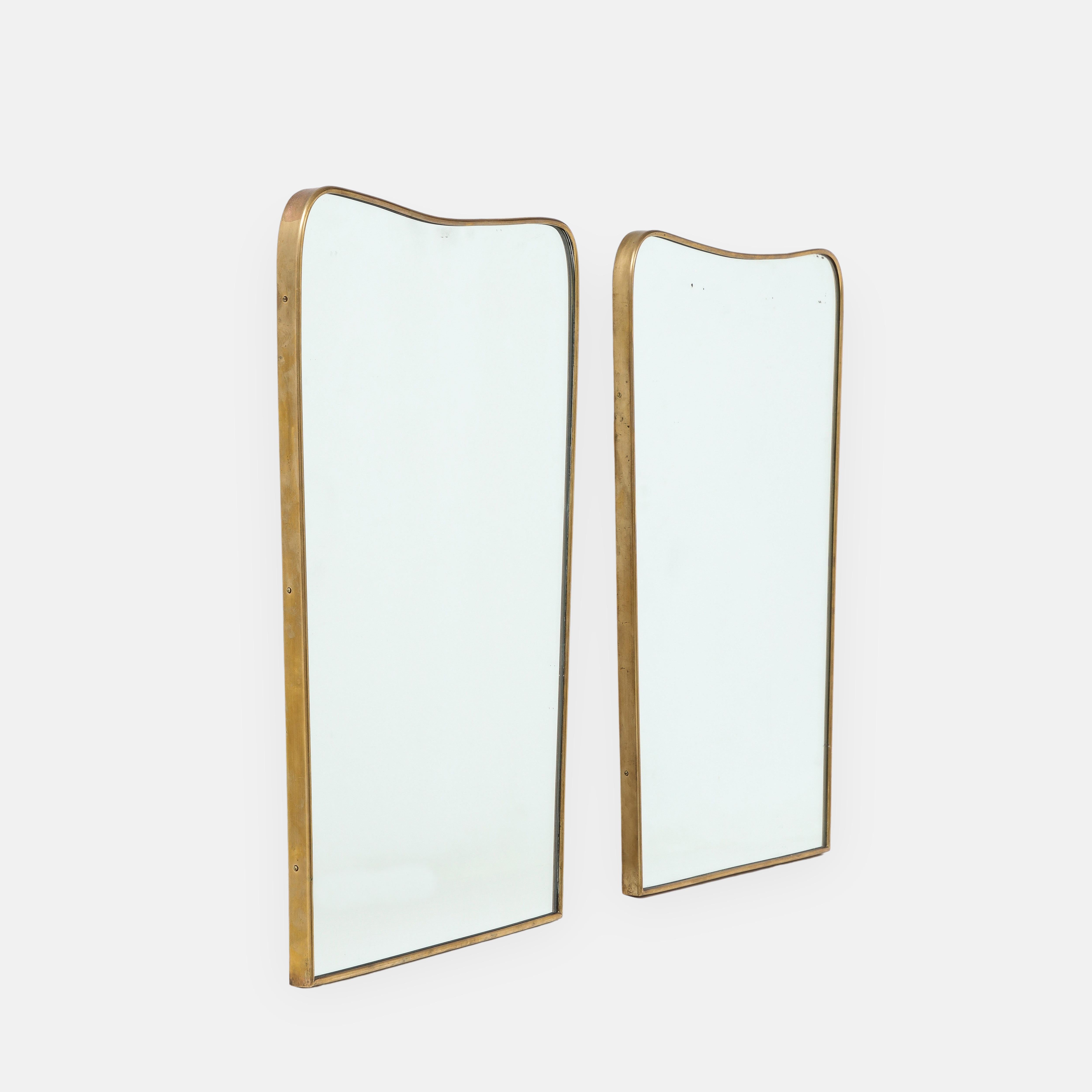 1950s Italian modernist pair of wall mirrors consisting of shaped brass frames with gently arched tops and rounded corners which taper towards the bottom with wood backings. Their original mirrored plates show some areas of foxing which illustrate