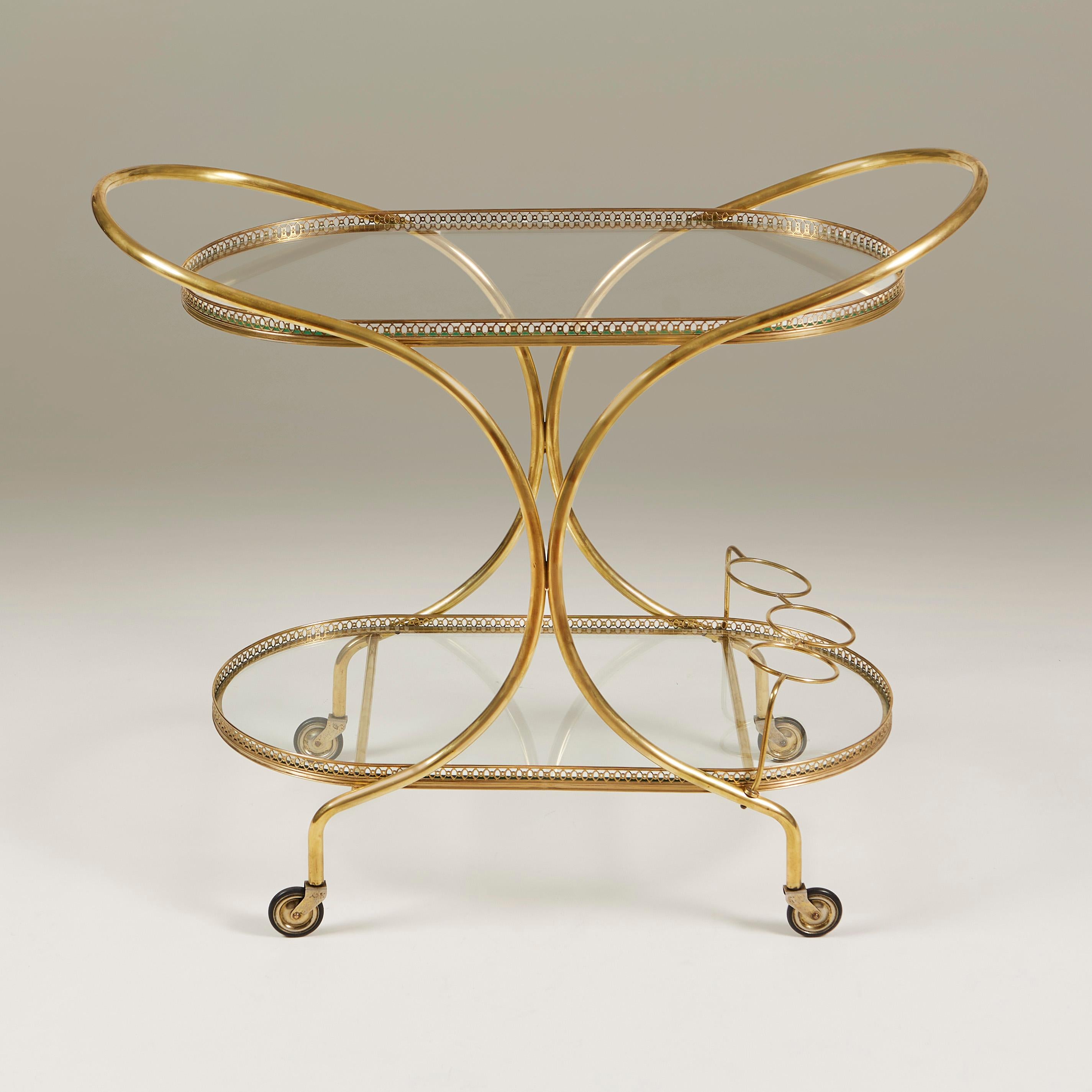 Elegant two-tier oval drinks trolley with simple curved brass frame that extends to create a handle either end. Decorative filigree brass edge to both shelves. Simple brass bottle holder for 3 bottles. Sits on castor wheels for ease of movement.
