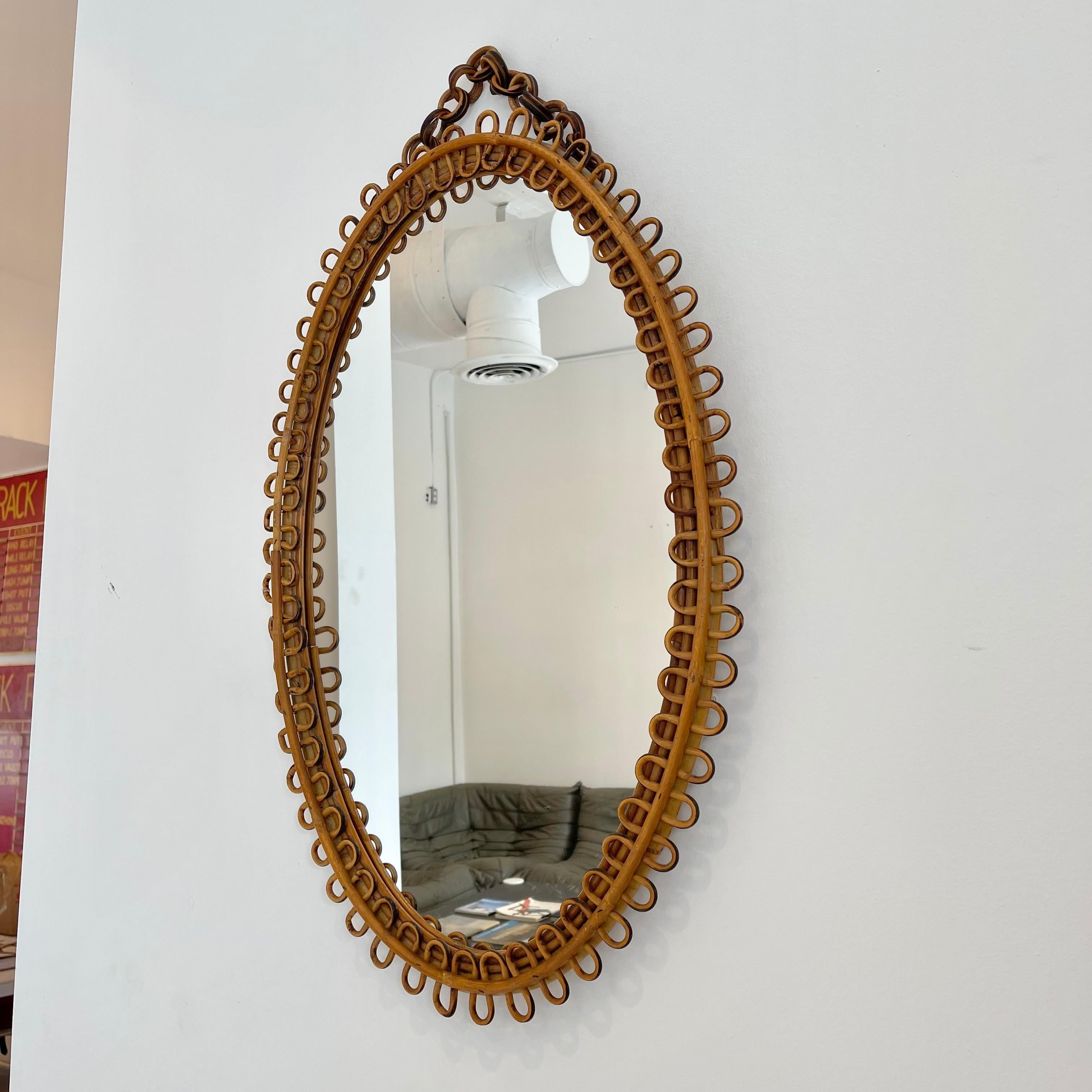 Gorgeous oval rattan hanging mirror from Italy, circa 1950. A single reed of rattan which bends in a bow-like manor adorns the face of the frame. This reed is fastened to a solid rattan frame. Mirror hangs from a chain of wood rings. Good vintage