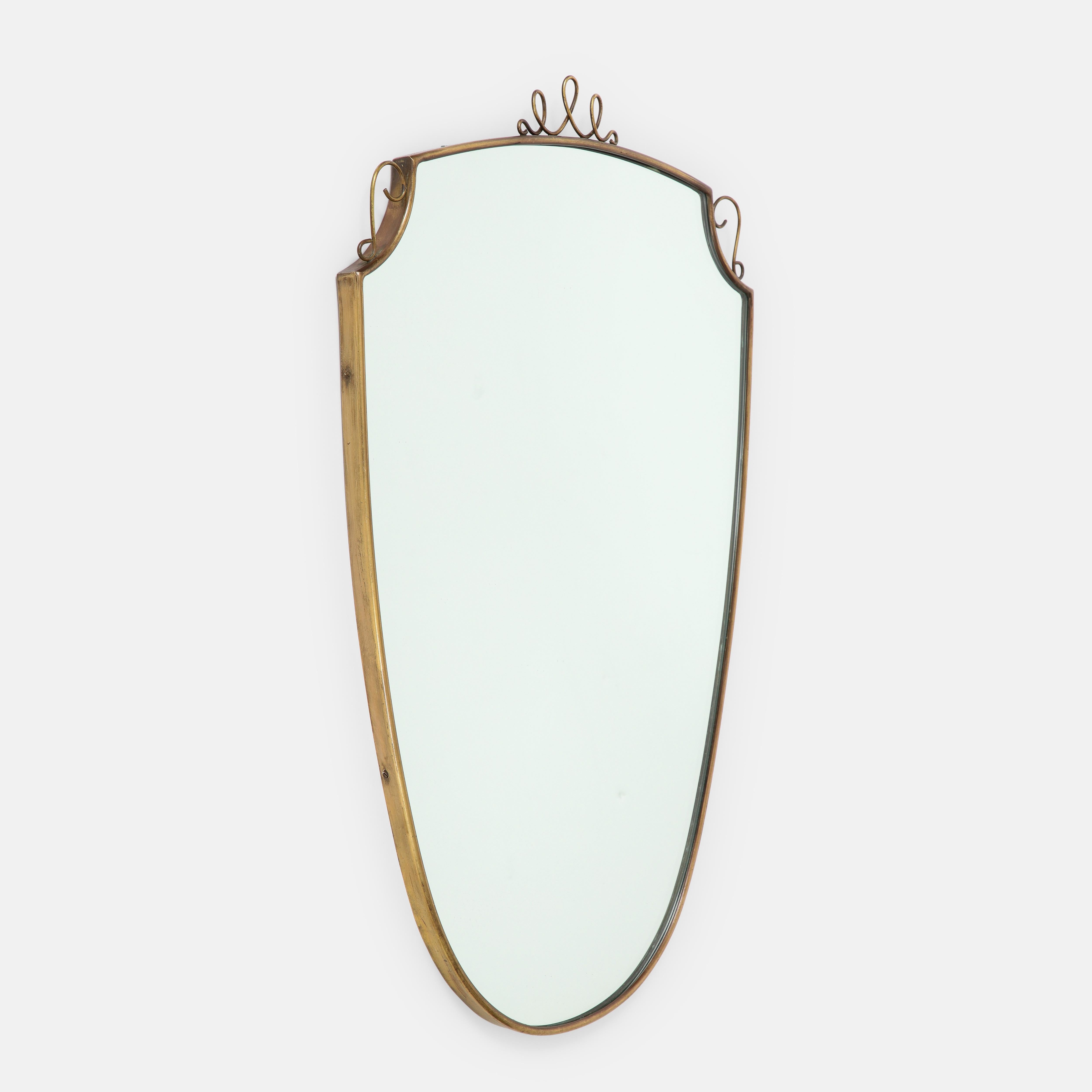 1950s Italian shield shaped brass wall mirror with scroll decorations. This original midcentury period mirror has beautiful rich patina to the brass with wood backing.