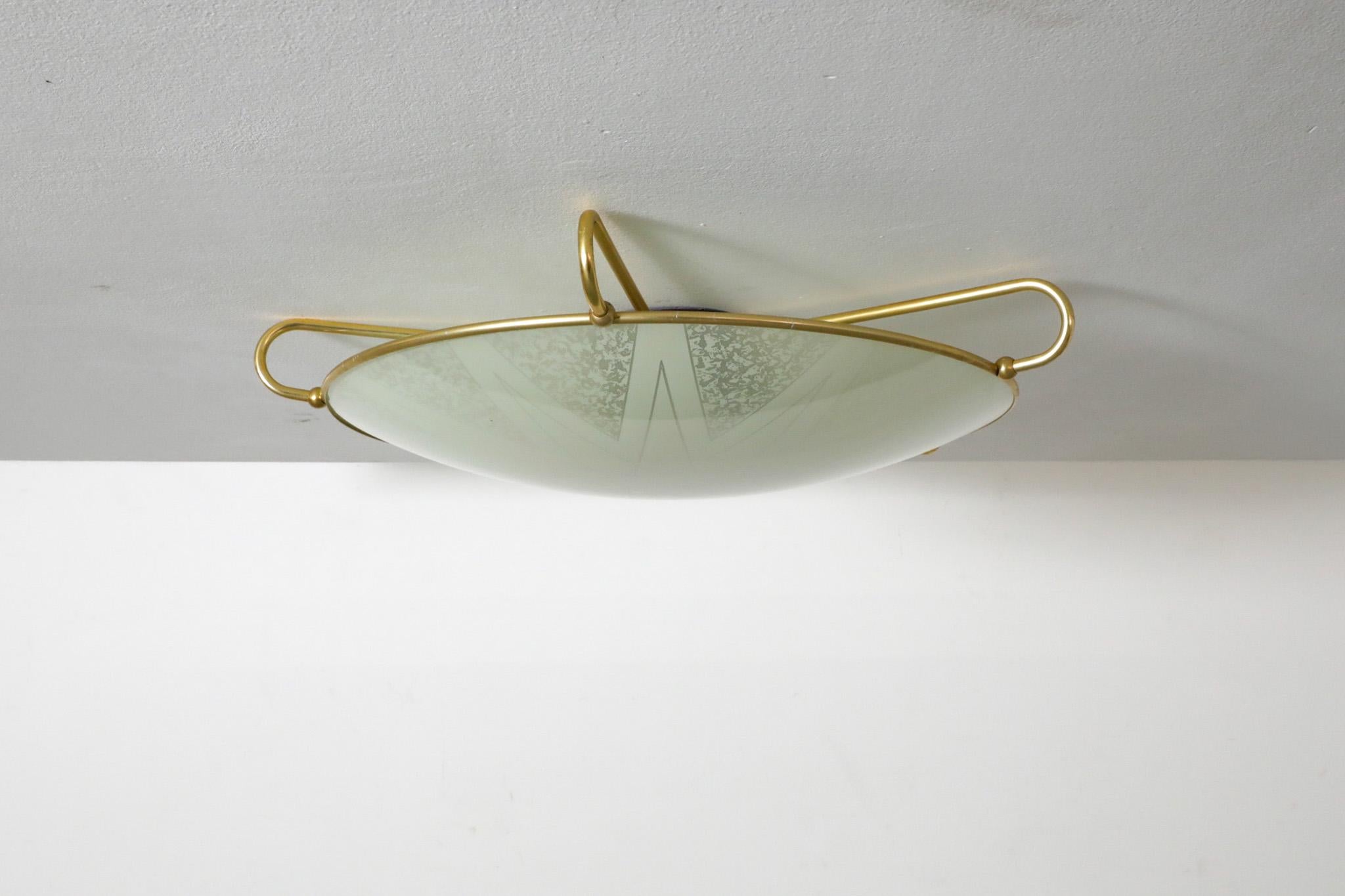 Flush mount ceiling or wall lamp with decorative glass rondelle by Erco. Bakelite base and brass toned arms that hold the disc in place. The glass dish sits inside polished brass toned arm. The light takes 3 medium base bulbs; LED recommended due to