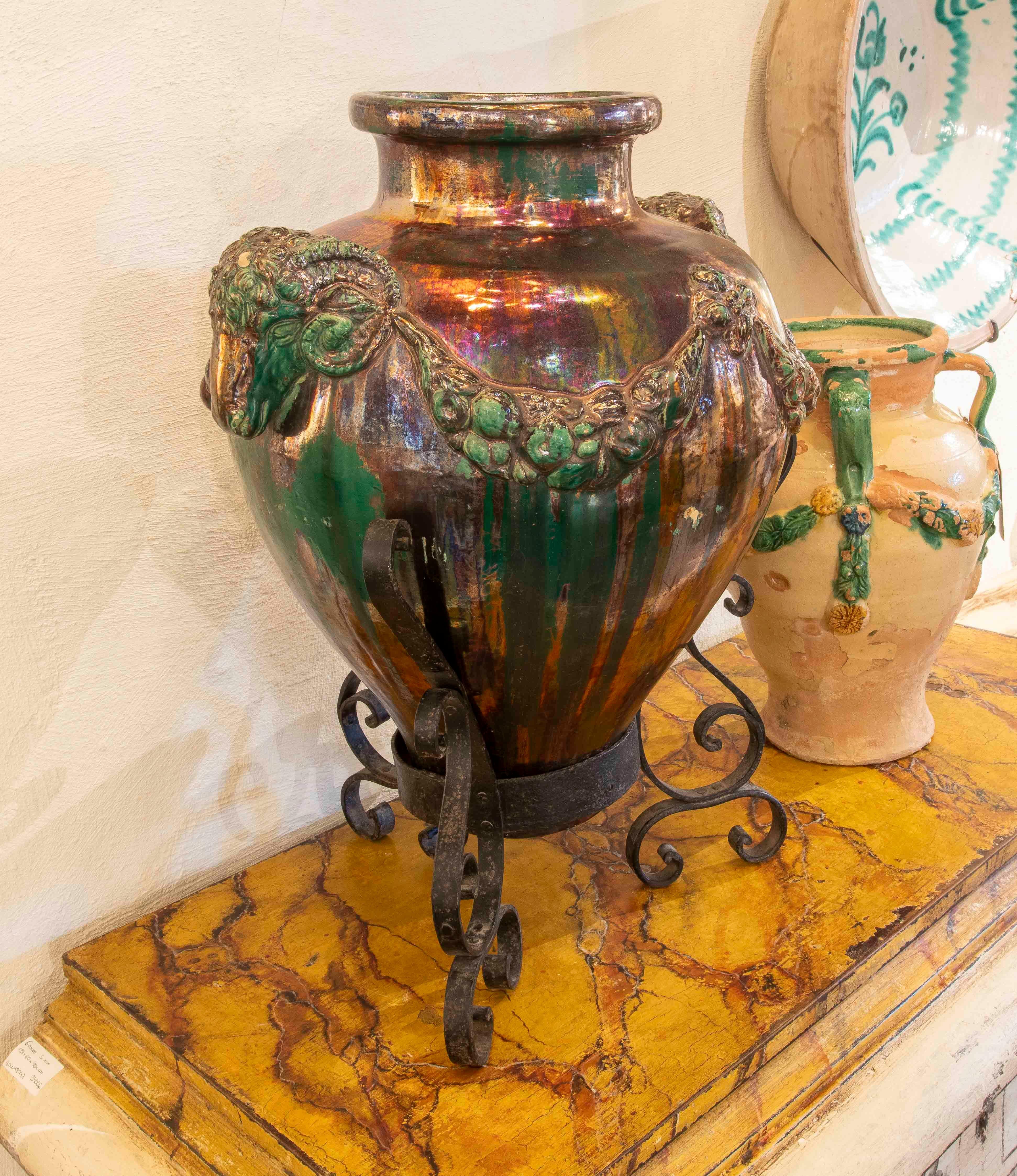 A stunning large glazed ceramic vase with an iridescent finish in shades of green, ochre, and copper. The piece features a relief decoration of rams' heads linked by garlands of fruit, giving it a classic and sculptural feel.

The vase rests on a