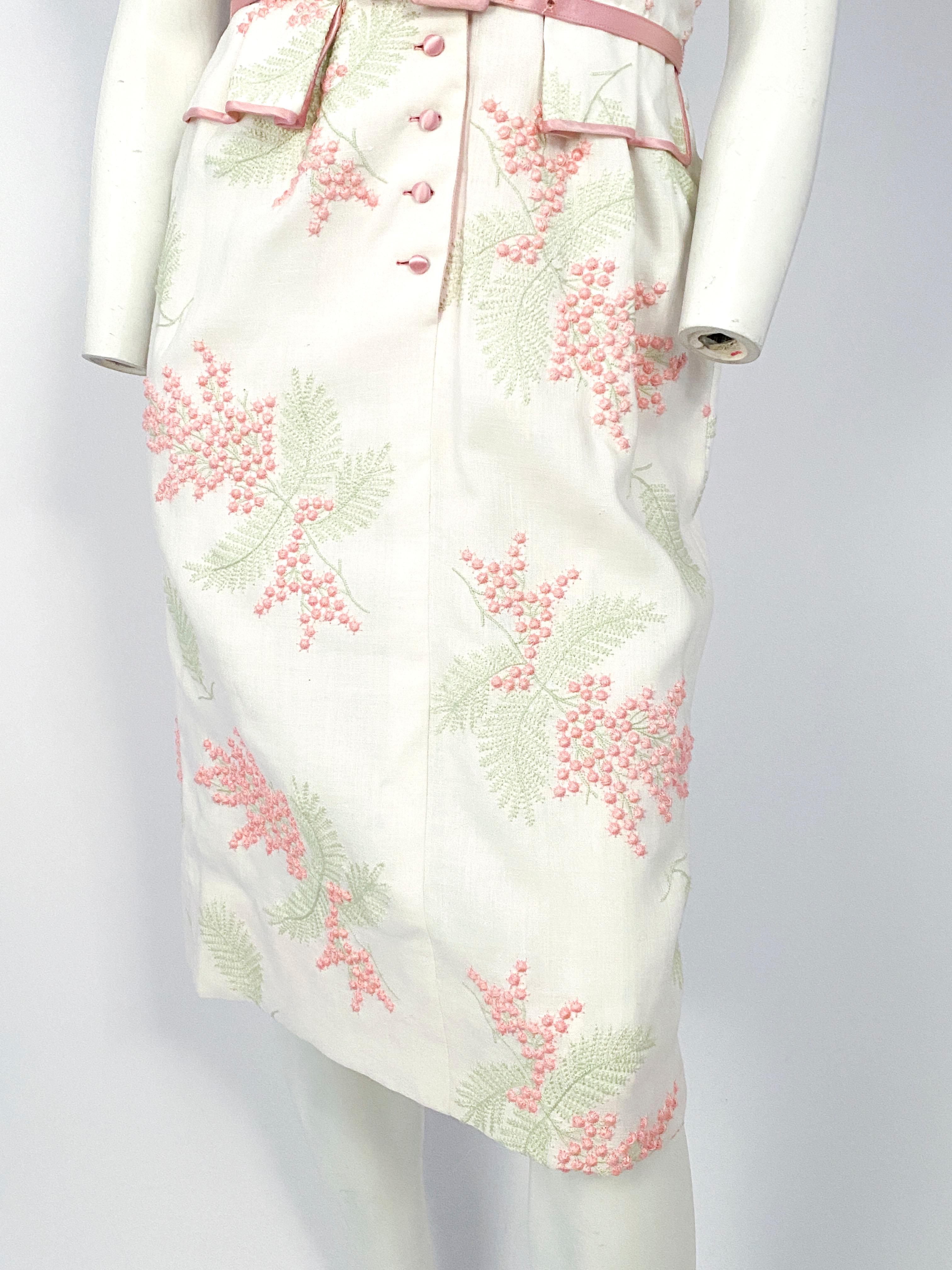 1950s white linen day dress with pink and green raised floral and foliage embroidery, satin covered buttons, piping, lapel, and matching satin belt. Short sleeves are finished with a wide cuff and waist is cinched with faux pocket covers.