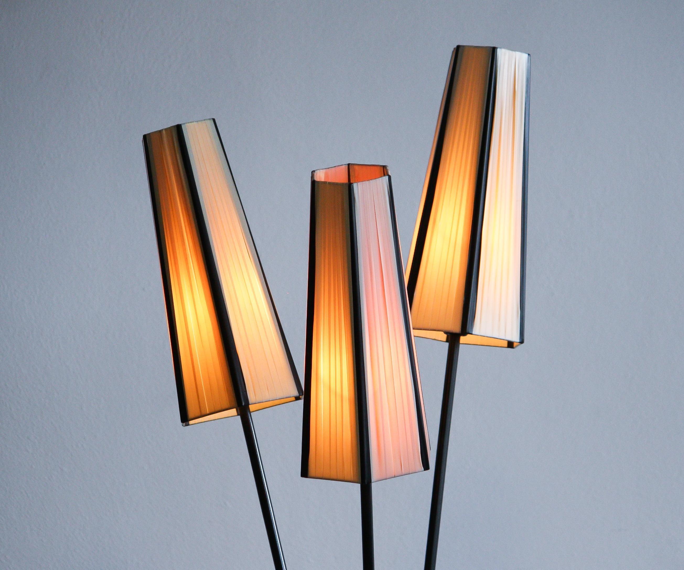 Floor lamp with two off-white and one salmon polyester fabrics covers from the 1950s.
The caps are lined with black cords.
The lamp stands on a black metal three-leg rest standard and the caps on brass rods. It is in good condition.
Period