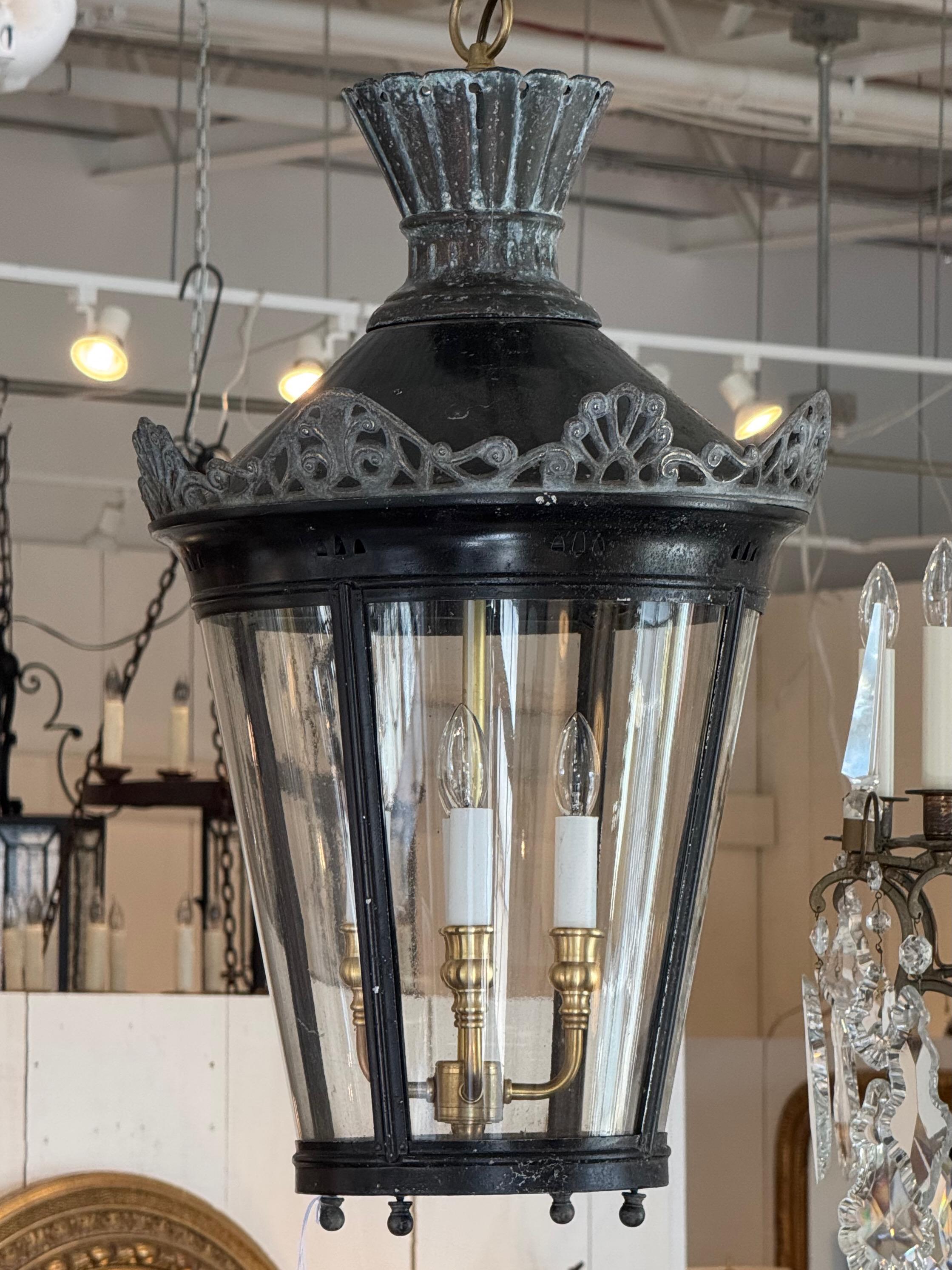 Large metal lantern with a tapered glass-paneled frame and pierced foliate detailing along the rim. The patinated black finish contrasts with the brass three-light interior, creating a timeless balance of industrial and classical elements. Suspended