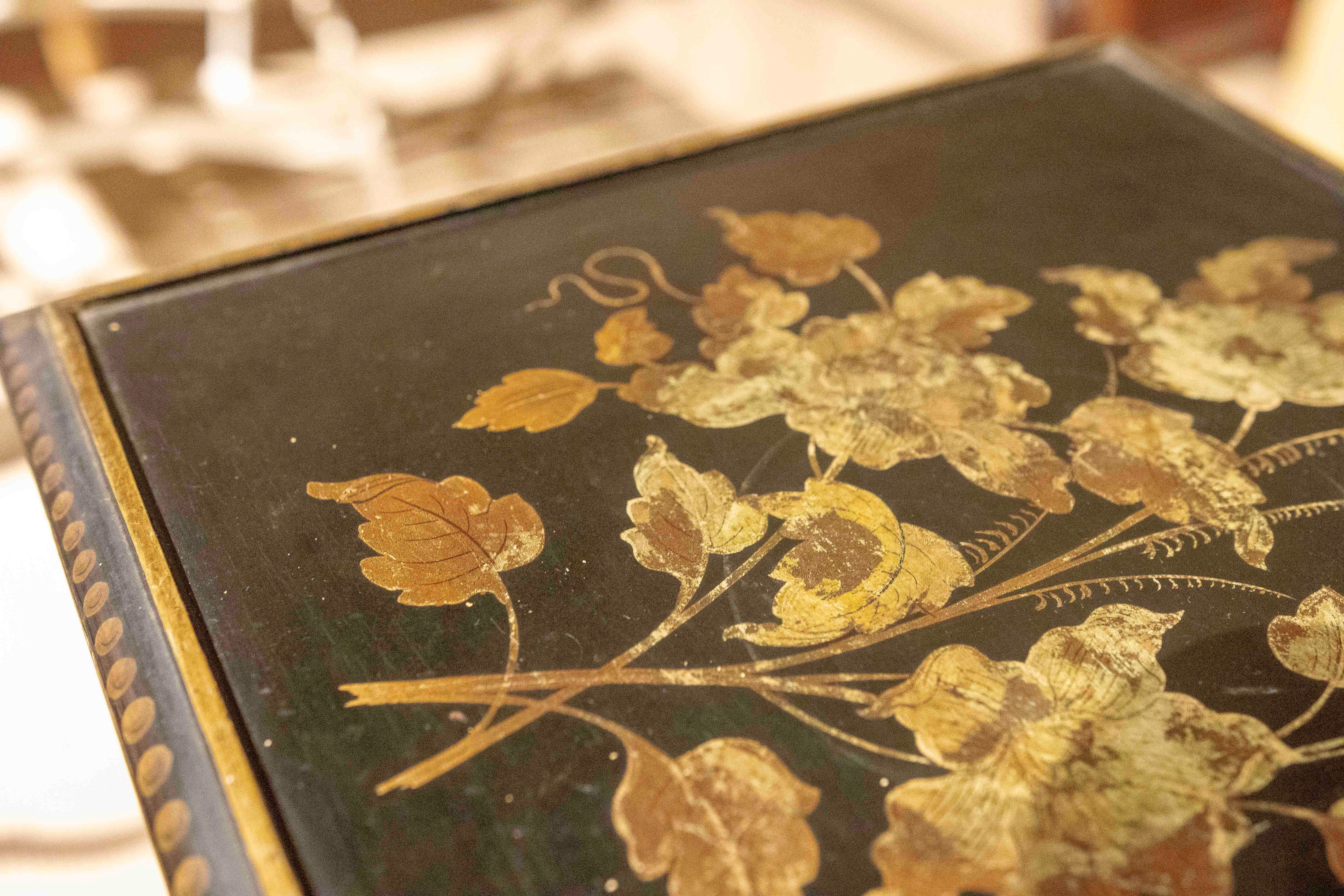 Élégante table d'appoint en métal à la structure fine et raffinée en noir. Son enveloppe carrée présente une délicate décoration florale peinte à la main dans des tons dorés et bruns, rehaussant son style vintage et sophistiqué. Le bord de la