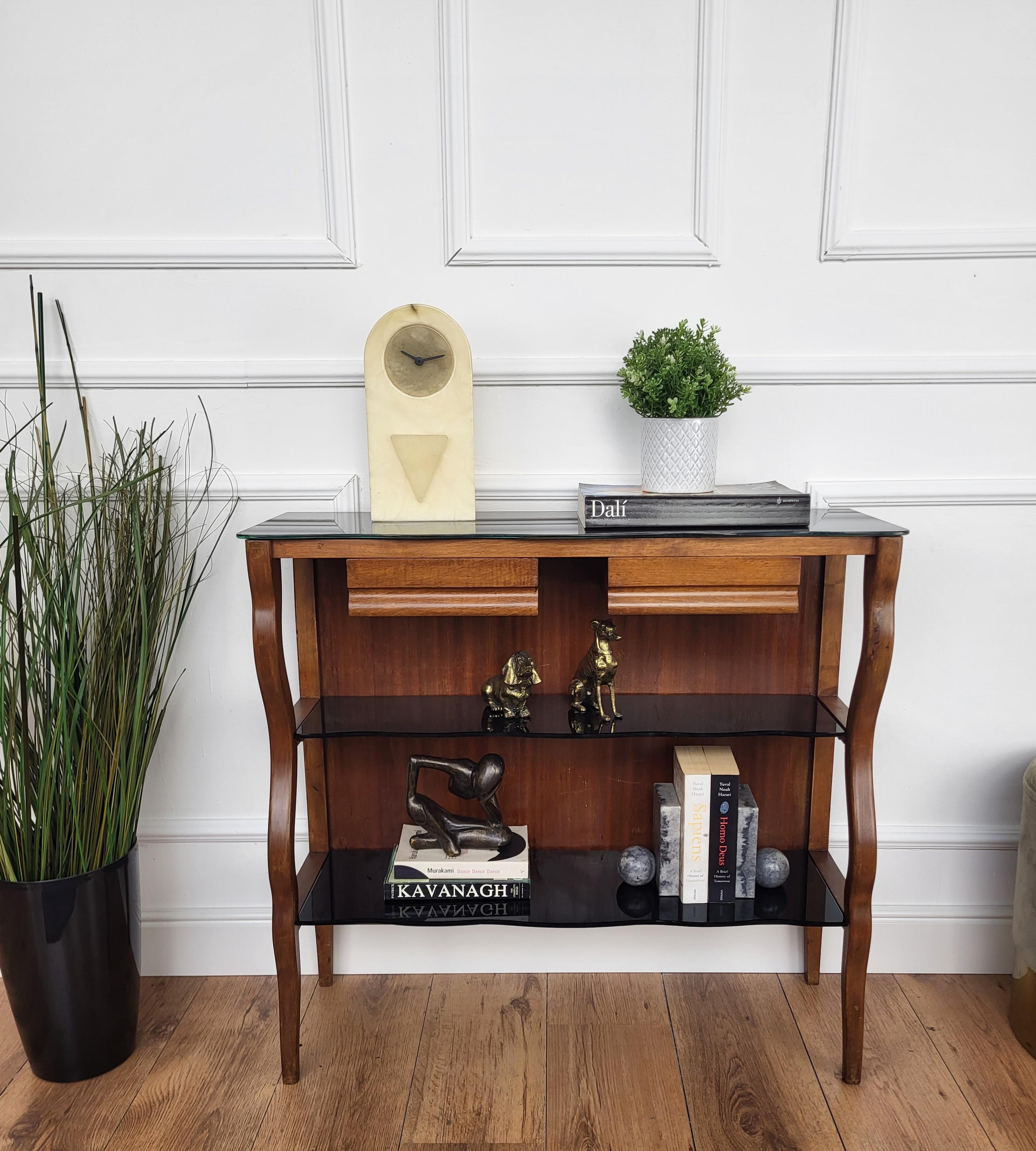 Très élégante et raffinée table console italienne des années 1950 de style Mid-Century Modern et Art Deco. Reposant sur de magnifiques pieds en bois, elle est mise en valeur par trois étagères en verre noir au décor arrondi et complétée par deux