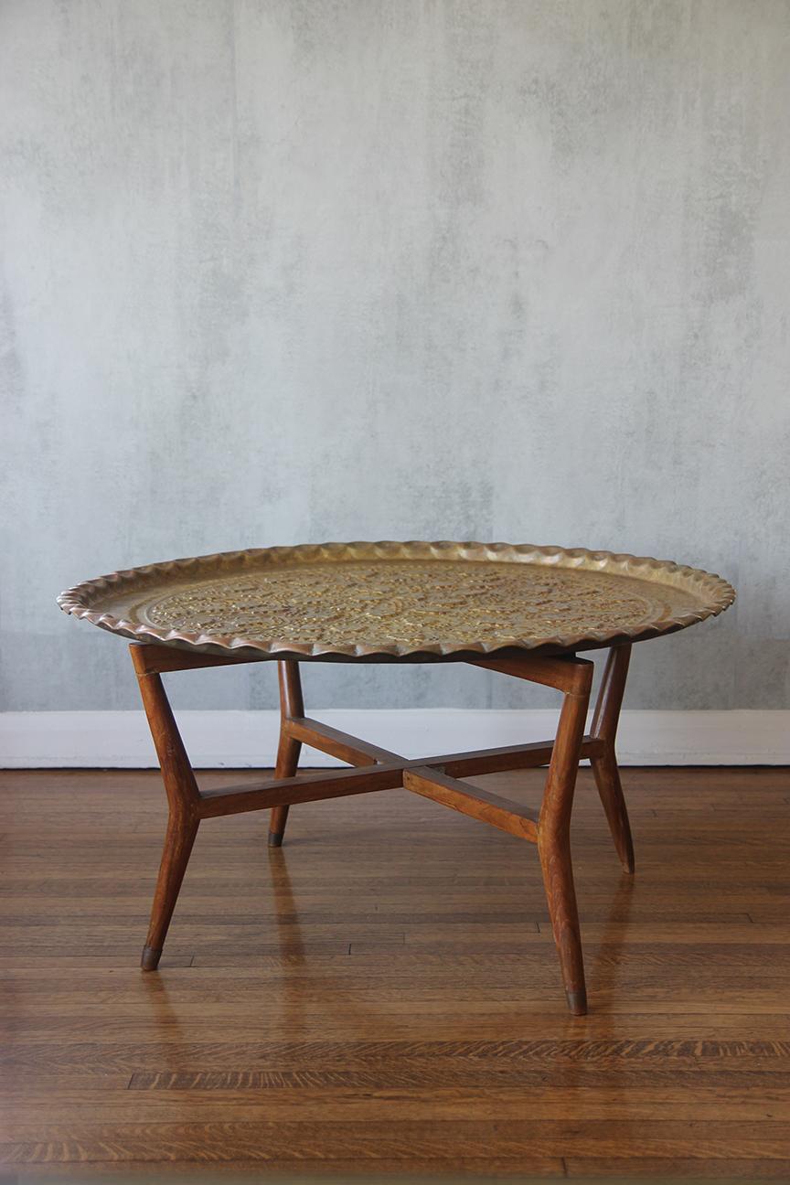 This exquisite hand-hammered brass Moroccan tray features a stunning mid-century mahogany base complemented by four elegantly designed folding legs. Versatile in its use, the piece can function as a stylish table for gatherings or be folded neatly