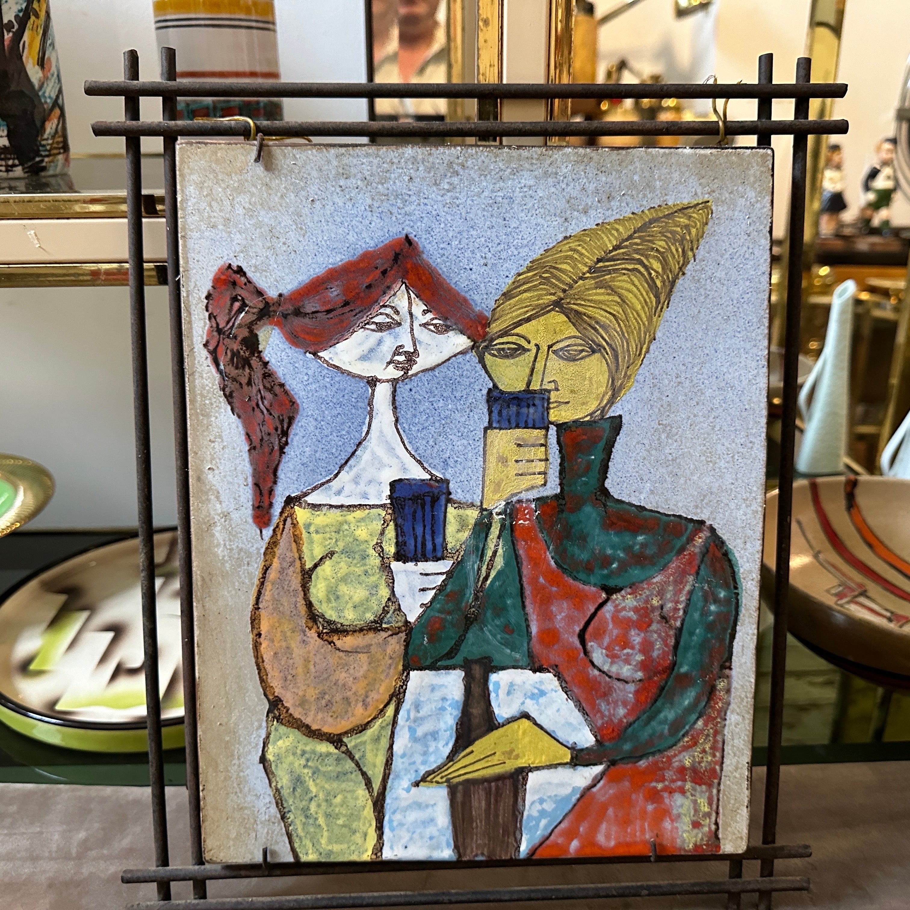 This 1950s Mid-Century Modern Italian ceramic plaque, framed in iron, is a vibrant and expressive example of the era's love for both abstraction and craftsmanship. The plaque features a hand-painted design depicting two stylized figures, a man and a