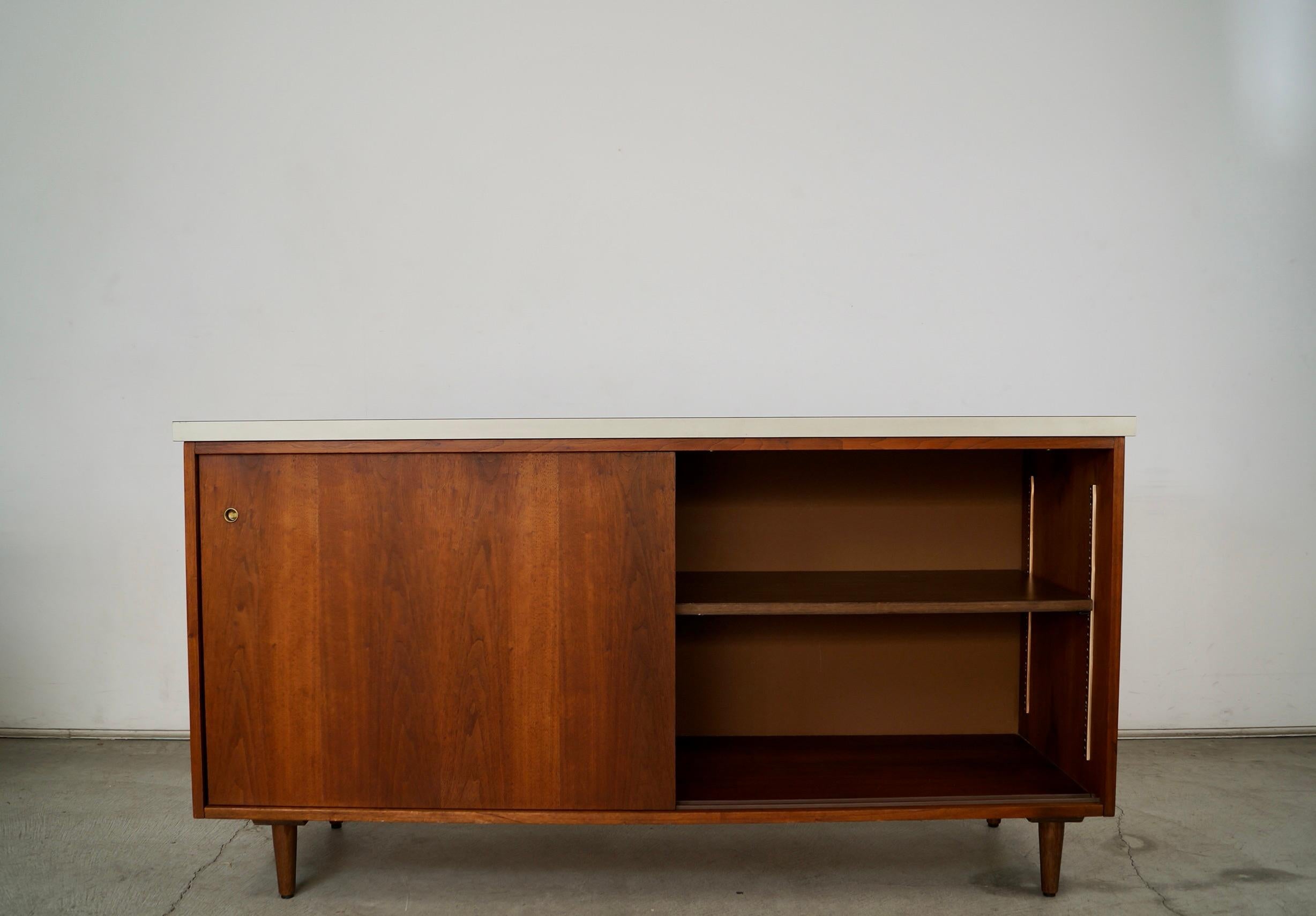 Se vende aparador de puertas correderas original de la época Moderna de mediados de siglo. Tiene dos puertas correderas que se abren a un estante ajustable a ambos lados, y está muy bien hecha. Es de nogal con la parte superior de formica blanca, y