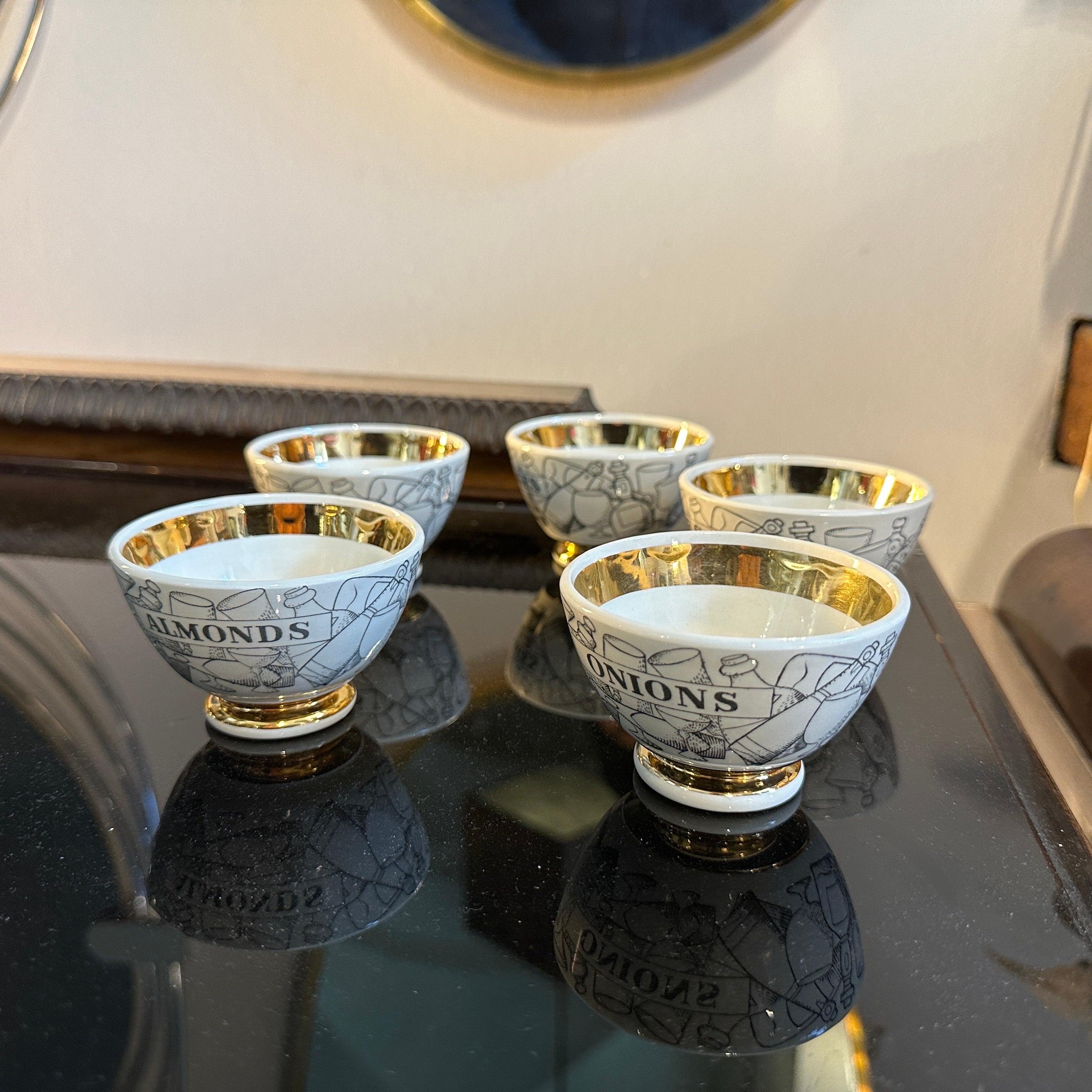 A set of five vintage aperitivo bowls by Fornasetti. These bowls feature intricate black and white illustrations with gold accents, especially on the rims and bases. Each bowl is adorned with text, likely labeling different types of aperitifs or